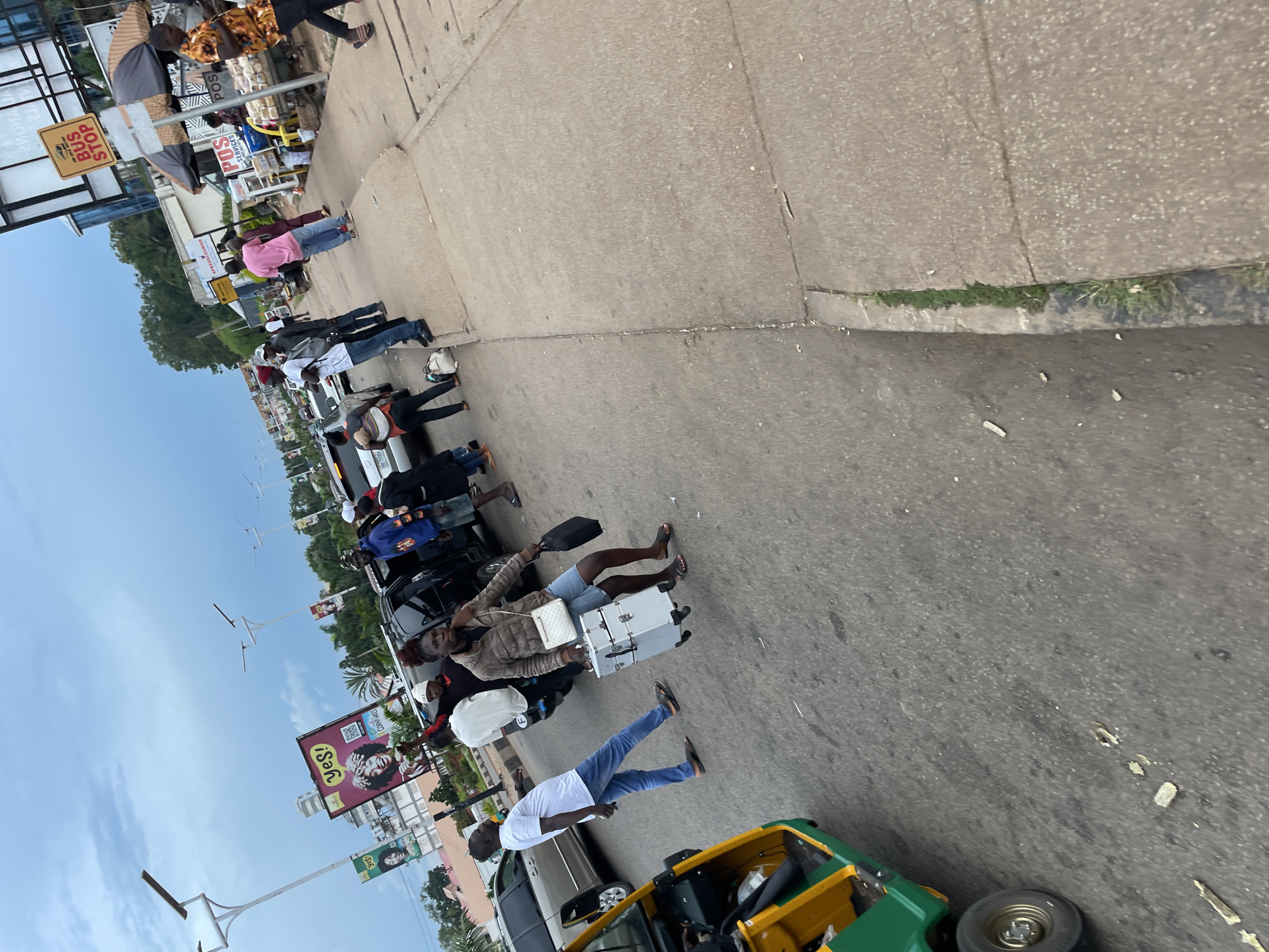A street scene in Jos, Nigeria, features a group of people gathered around a street, with several vehicles and structures visible. In the foreground, a woman is carrying a large, silver, wheeled suitcase, and a black bag. She is wearing a brown jacket and denim shorts. To her left, a man in a white t-shirt and blue jeans walks away from the camera. A green and yellow three-wheeled vehicle is parked on the left side of the road.
Further back, a black SUV is parked on the street, with several people standing near it. One person, wearing a blue hooded sweatshirt, is leaning into the open driver's side door. A few individuals are visible on the sidewalk, including a person in a pink shirt. Behind the people on the sidewalk, there are market stalls and buildings, including one with a "BUS STOP" sign. In the background, there are trees and a cloudy sky. Several billboards are also visible. The overall environment appears to be a busy urban area.