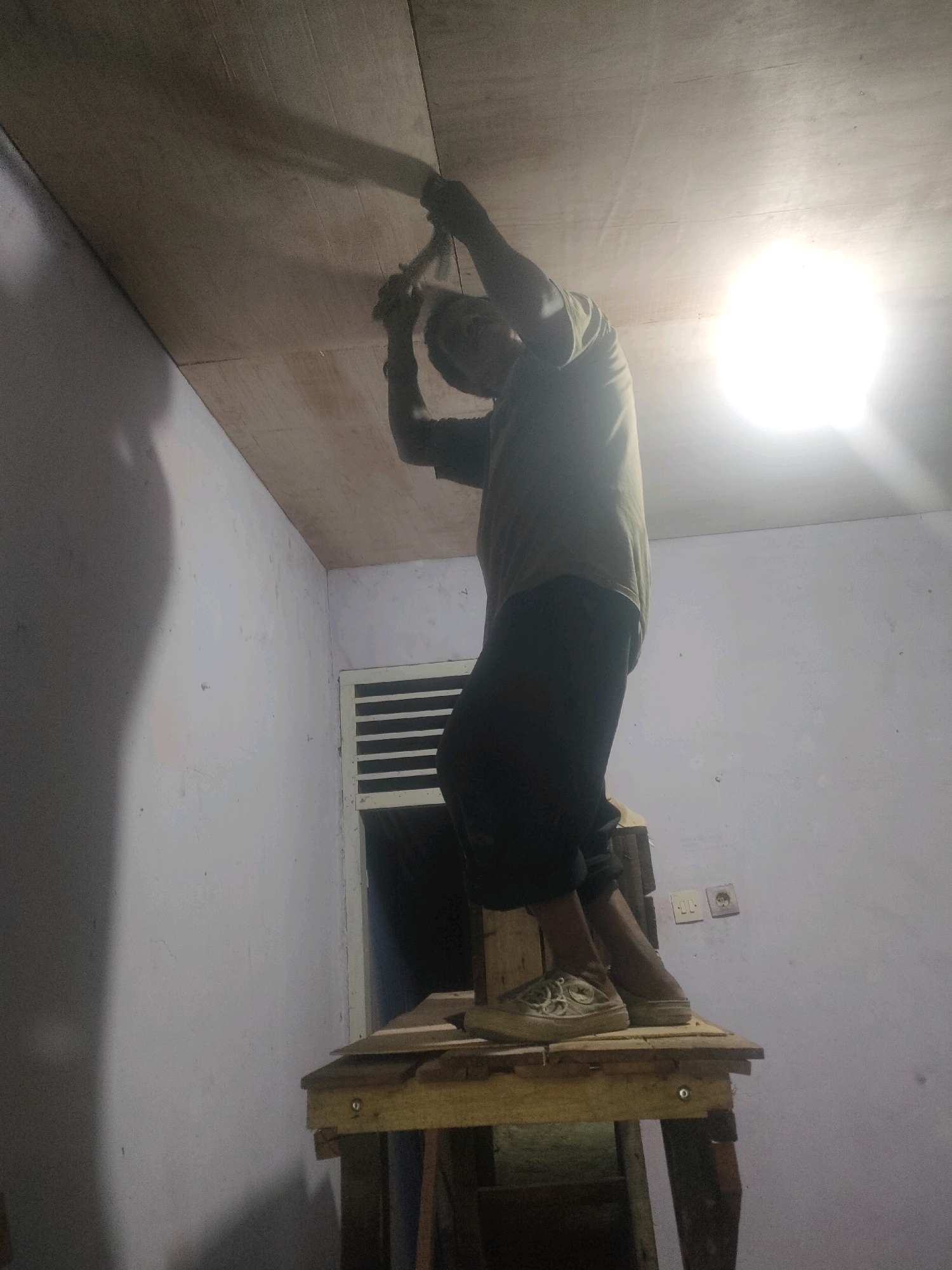 In Pekanbaru, Indonesia, a person stands on a makeshift wooden platform, appearing to be engaged in work on the ceiling. The person is wearing a dark t-shirt and dark pants, with light-colored shoes. They are holding a tool, possibly a hammer or a similar implement, and are positioned beneath a wooden-paneled ceiling. The room is sparsely furnished, with a white wall to the left casting a shadow. To the right, a window with horizontal blinds is visible, along with electrical outlets on the wall. A bright light source, possibly a lamp, illuminates the scene from the upper right. The overall impression is of a domestic or unfinished construction environment.