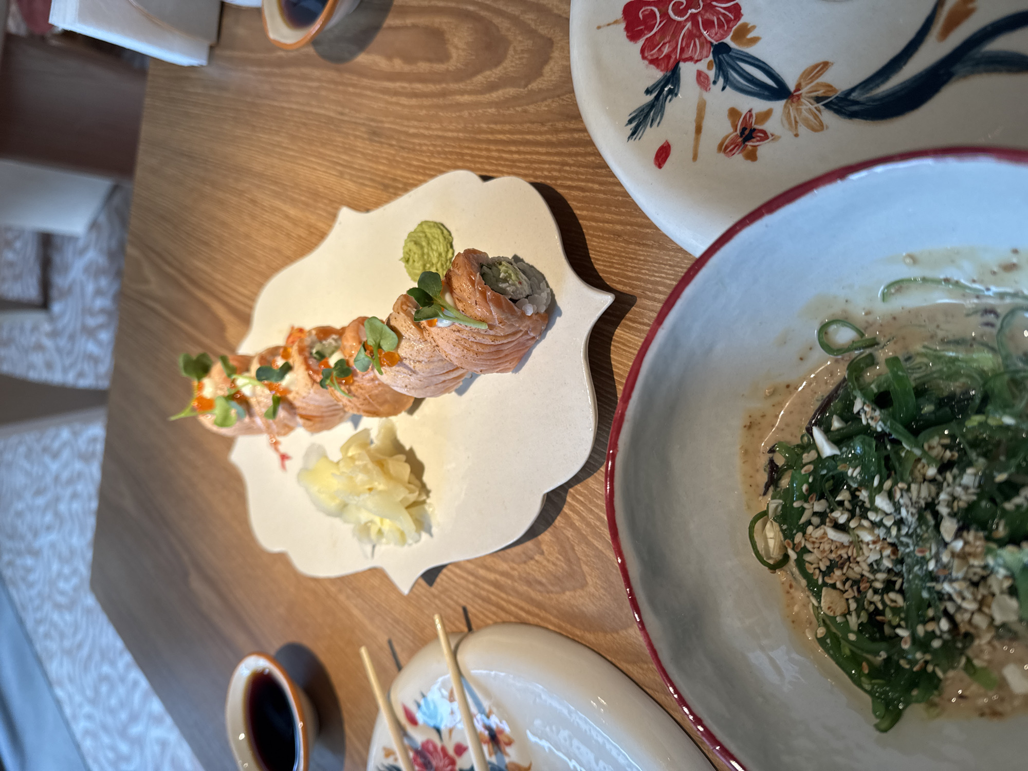 On a wooden surface, possibly a table, is an assortment of dishes. A decorative, flower-patterned plate sits near a bowl filled with green seaweed salad sprinkled with sesame seeds and immersed in a light-colored sauce. Adjacent is a plate of salmon sushi rolls, accompanied by wasabi and pickled ginger. Nearby, a small dish of soy sauce and chopsticks suggest a dining experience. The location is unknown.