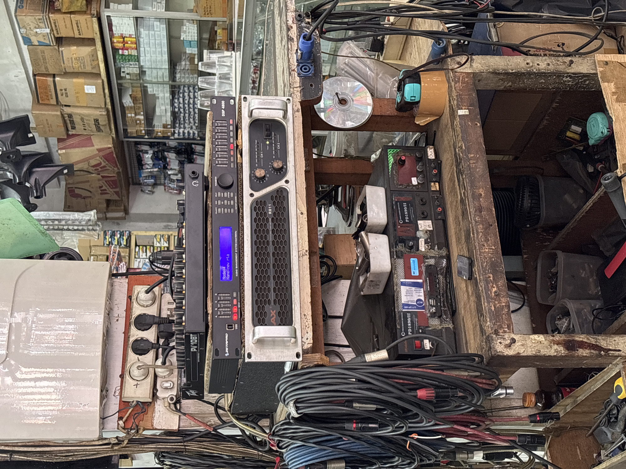 The image depicts a cluttered audio equipment shop in Denpasar, Indonesia. The foreground is dominated by various audio devices stacked on wooden shelves. From top to bottom, a PV10BT mixer is visible, followed by a DSP 4800 audio management system, and an R811 professional power amplifier. Numerous audio cables, including XLR and speaker cables, are coiled and hang around the equipment. A small, old-fashioned power strip with multiple outlets is mounted on a white structure above the mixer.
The middle ground shows more audio equipment, specifically a Precision Devices PD1890/II unit and an RCF Audio Console. A roll of brown tape and a teal-colored measuring tape are placed on the shelf near these devices. A CD is also present on the shelf.
The background reveals shelves filled with boxes and some transparent plastic cups. A glass display cabinet is also visible, containing various electronic components and accessories. The overall environment suggests a busy, operational retail space focused on audio equipment. There are no people visible in the frame. The lighting appears to be a mix of ambient and possibly artificial light, casting shadows and highlighting the textures of the wooden shelves and equipment.