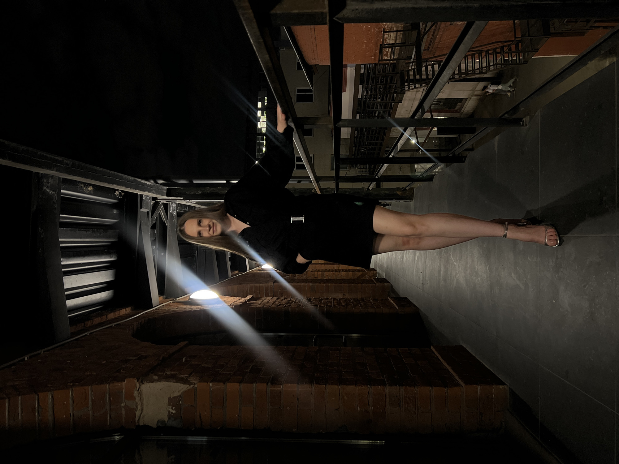 A woman in a black dress is standing in an outdoor corridor. She is holding onto a dark metal railing with one hand. Behind her is a brick wall with lights on it. Across the corridor are fire escapes and a taller building. The scene appears to be in Moscow, Russia.