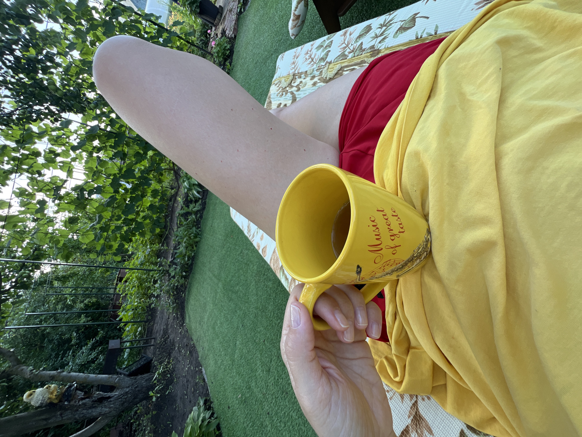 In Lopatino, Russia, a person rests on a lounge chair in a garden, holding a yellow mug with "Music of great taste" written on it. The mug contains a dark liquid, likely tea. The person is wearing a yellow top and red bottoms. The background features lush green foliage, a tree with a decorative owl, and a grill. Artificial turf covers part of the ground. The scene is bathed in natural light, suggesting it is daytime.