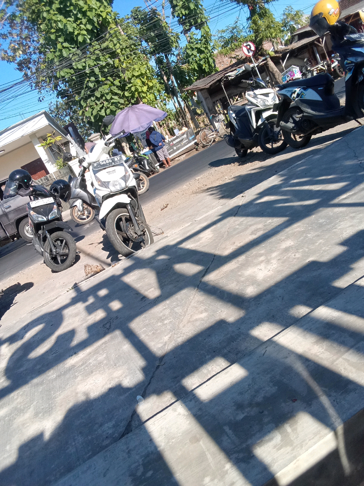 The image depicts a street scene in Jember, Indonesia, characterized by the presence of several parked motorcycles. The foreground is dominated by the shadow of an overhead structure, casting geometric patterns on the concrete pavement. Two black motorcycles, each with a helmet placed on its seat, are parked on the left side of the road. A white motorcycle with a blue and white design is parked further down the street, also with a helmet on its seat. In the background, a few individuals are visible near a small roadside structure, with a purple umbrella providing shade. The environment appears to be a sunny day with clear blue skies, and lush green trees line the street. The overall impression is a typical urban street scene in Indonesia, with motorcycles being a primary mode of transportation.