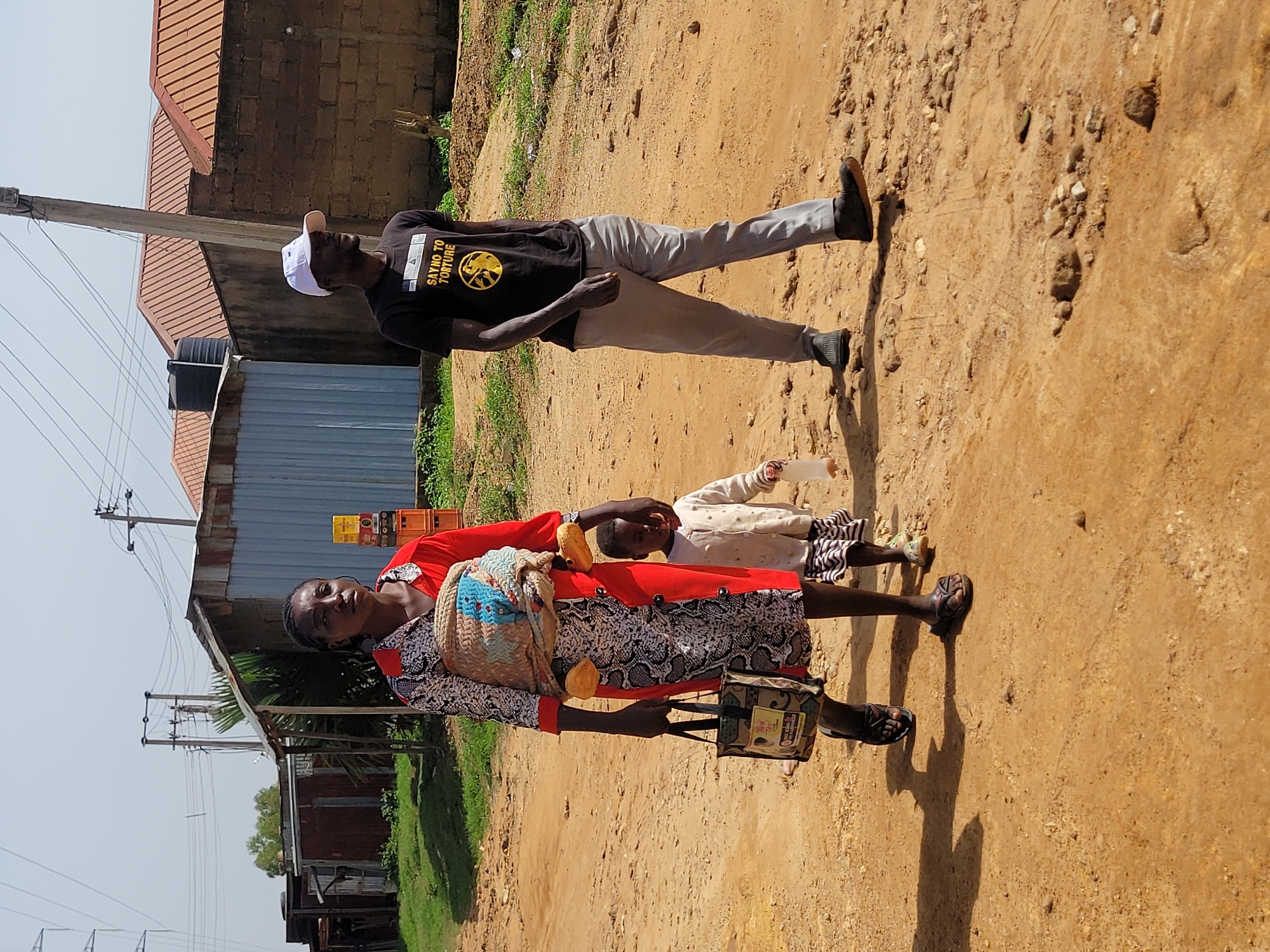 In a dusty, outdoor environment in the city of Jos, Nigeria, a woman, a young child, and a man are walking on a dirt path. The woman is in the foreground, wearing a red and white patterned dress and carrying a woven basket on her back, secured with fabric. She also carries a dark-colored bag in her left hand. The child, who appears to be a toddler, is walking beside the woman on her right, wearing a light-colored knitted sweater and striped skirt, and holding a white plastic bottle. To the right of the woman and child, the man is walking, dressed in a black t-shirt with yellow text that reads "SAY NO TO TORTURE", and light grey trousers. He is wearing a white baseball cap and dark shoes. The background shows a few buildings with corrugated metal and brick construction, some with red-tiled roofs. Power lines and poles are visible overhead, along with some vegetation and a water tank. The ground is a mixture of dirt and small rocks, with some shadows cast by the subjects and overhead structures. The lighting suggests it is daytime, likely with bright sunlight.