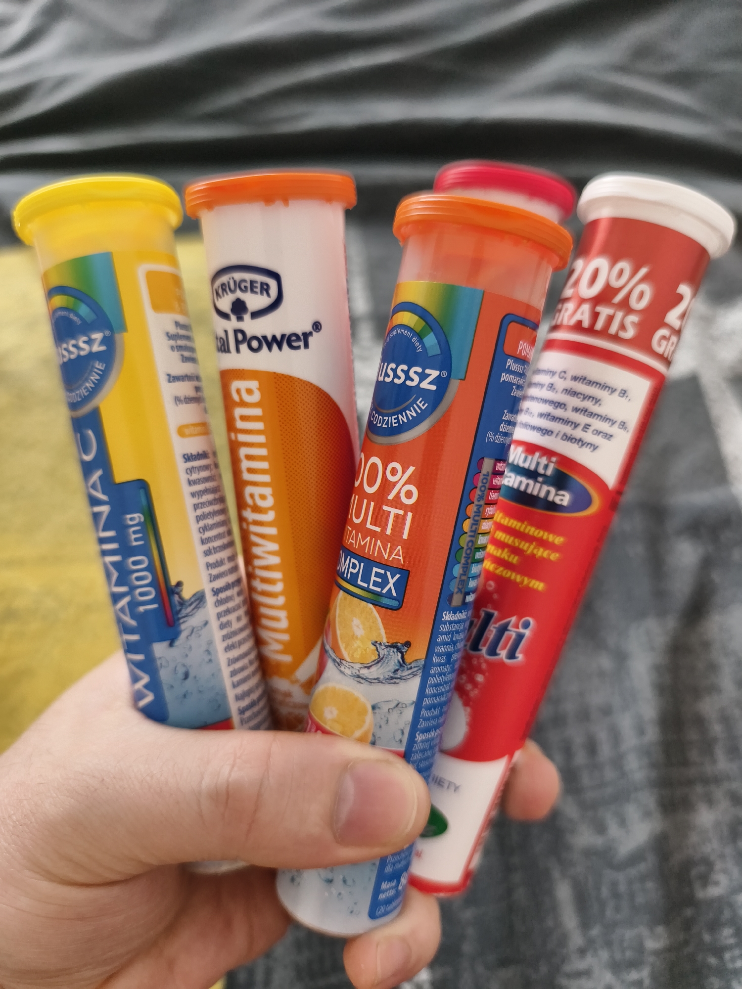 Four tubes of effervescent tablets are held in a person's hand against a dark, textured background, possibly a blanket. From left to right, the tubes are yellow, white with orange text, orange with white text, and red with white text.
The yellow tube prominently displays "WITAMINA C 1000 mg" and a circular blue logo with the letters "USSSZ" and "CODZIENNIE". The white tube with orange text reads "KRÜGER" and "tal Power®". The orange tube, slightly obscured, has "100% MULTI TAMINA COMPLEX" and an image of an orange slice with water. The red tube has "20% GRATIS GR" and "Multi" in large white letters.
The background appears to be a dark gray, possibly a fabric with a subtle pattern, suggesting an indoor setting. The lighting is somewhat soft, creating subtle shadows on the tubes. The hand holding the tubes is detailed, showing skin texture and fingernails. There is no discernible urban environment or specific landmarks visible, so the location in Tczew, Poland cannot be determined from the image.