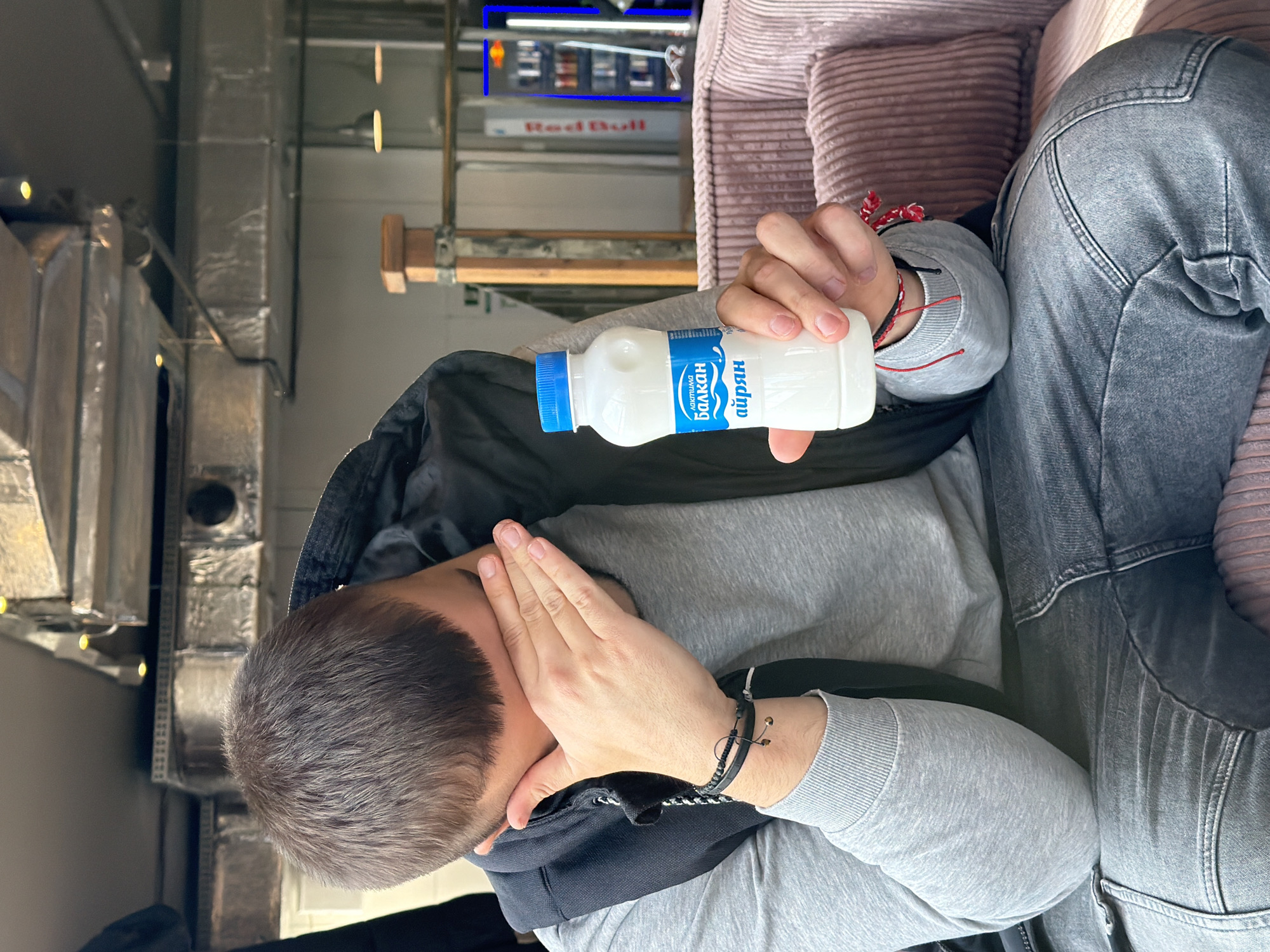 A man with short brown hair sits on a pink corduroy couch, head in his hand in an apparent gesture of despondency, while holding a small white plastic bottle of "Балкан Айрян" in his other hand. He wears a gray sweatshirt and black jacket. He is wearing a black beaded bracelet. He sits next to a person wearing gray denim pants. A "Red Bull" neon sign is visible in the background. Based on the product and architecture visible, the location is plausibly in Sofia, Bulgaria.