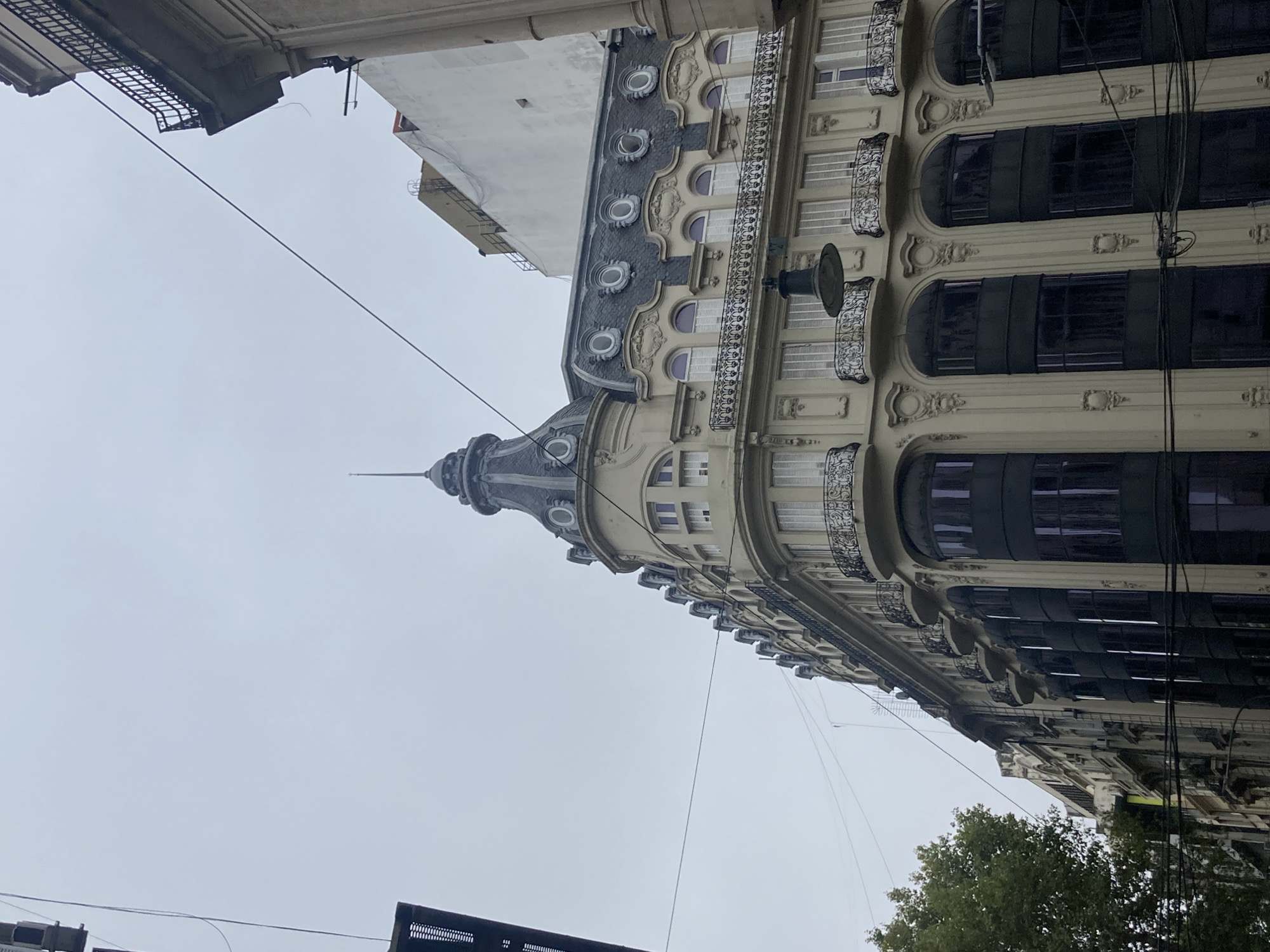 The image presents a low-angle view of an ornate, multi-story building against a uniformly grey, overcast sky. The building, likely situated in an urban environment, features a distinctive architectural style, possibly late 19th or early 20th century.
A prominent corner of the building rises into a conical spire, adding to its grand appearance. Below the spire, a dark, mansard-style roof is visible, punctuated by several decorative oval dormer windows with light-colored frames. The facade of the building is light-toned, adorned with intricate architectural details, including moldings and decorative frames around the windows. Multiple floors are discernible, each featuring balconies with elaborate black wrought-iron railings. Many windows are arched, while others are rectangular, and some appear to have dark, possibly reflective, glazing or blinds.
A notable feature is a large, dark, bell-shaped fixture or lamp projecting from the facade on one of the upper stories. Numerous black power lines or cables crisscross the frame against the sky and in front of the building, suggesting an older urban infrastructure. A small portion of green tree foliage is visible in the lower right corner, hinting at street-level vegetation.
The overcast conditions indicate daytime, but the diffused light suggests it could be an early morning, late afternoon, or simply a very cloudy day. There are no people visible in the image, nor is there any discernible text.