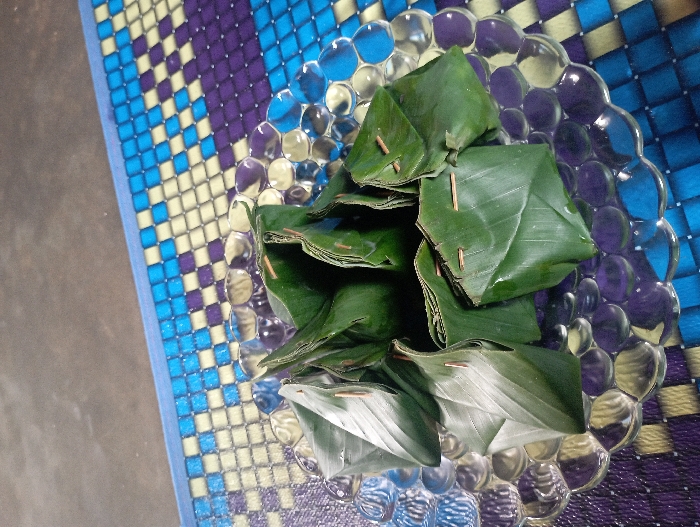 A clear glass plate, featuring a textured, bubble-like surface, holds a collection of individually wrapped food items. Each item is meticulously encased in a vibrant green banana leaf, folded into a triangular or pyramidal shape, and secured with small, slender wooden picks or skewers. The banana leaves appear fresh, suggesting a recent preparation. The plate reflects the colorful pattern of the mat it rests upon, creating distorted but visible squares of blue, purple, and light yellow.
The background is dominated by a woven mat with a distinctive checkered pattern. Bands of blue and light yellow squares run across the left side, transitioning to bands of purple and light yellow squares on the right side, underneath and around the plate. The weave of the mat is visible, indicating its texture.
The food items, likely traditional snacks or desserts from Indonesia, given the context of Pubian, are arranged casually on the plate, ready for consumption or display. The lighting is bright and even, highlighting the natural green of the banana leaves and the reflective qualities of the glass plate. No people are visible, nor is any specific activity taking place beyond the static presentation of the food. There is no visible text in the image.