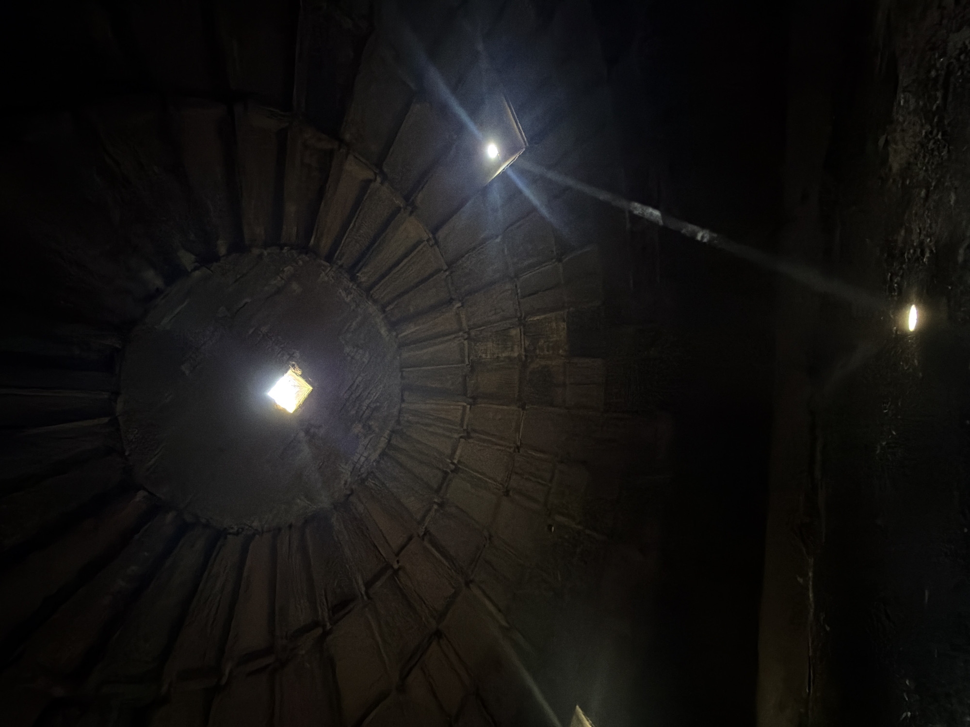 The image shows the interior of a dark, stone dome with a central opening at the top. Light streams in from the opening, illuminating the dome's interior structure composed of segmented, curved stone panels. A beam of light extends diagonally across the right side of the frame. The location is the Narikala Fortress in Tbilisi, Georgia.