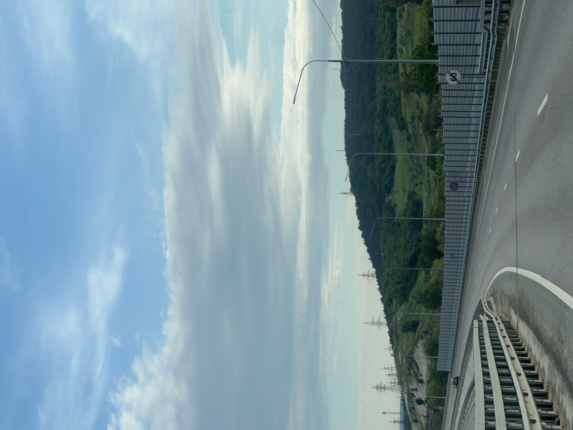 The image depicts a highway curving alongside a hillside with a mix of trees and grassy areas. The highway has a gray noise barrier along one side and a white line marking the edge of the lane. Streetlights line the road. In the background, high-voltage power lines are visible. A speed limit sign of 60 is posted. The sky is cloudy with patches of blue. The location is likely Inkerman, Ukraine.
