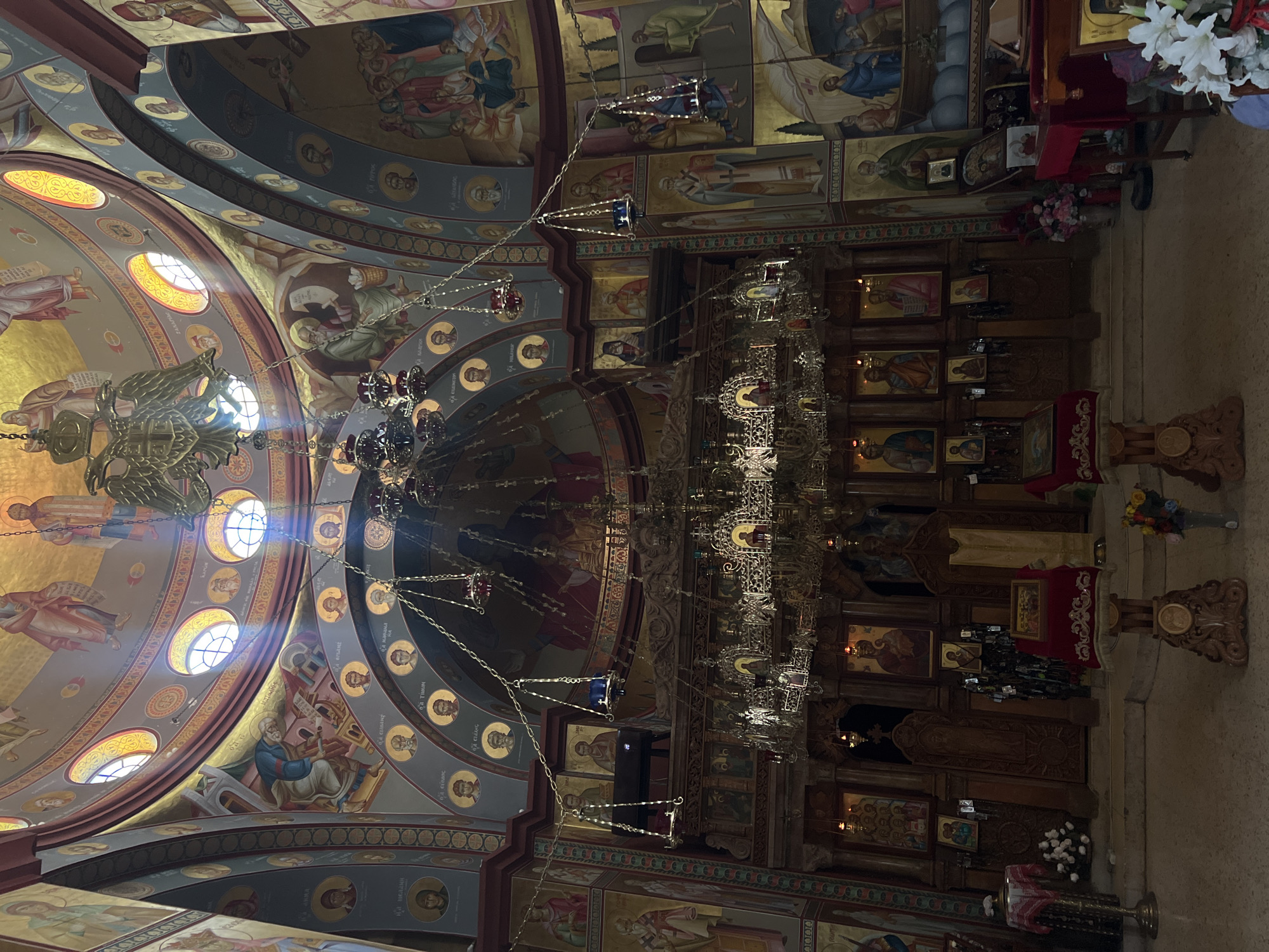The image depicts the interior of an Eastern Orthodox church in Emek HaYarden Regional Council, Israel. The scene is dominated by religious iconography, with extensive murals adorning the domed ceiling and walls. These murals feature depictions of saints and biblical scenes rendered in a traditional Byzantine style with gold leaf accents.
In the foreground, a large, ornate iconostasis, a decorated screen separating the altar from the nave, is prominently displayed. It is laden with icons of saints and religious figures, all framed by intricate woodwork. Hanging from the ceiling are several elaborate chandeliers and smaller votive lamps, some of which are lit, casting a warm glow. Sunbeams penetrate through arched windows in the dome, illuminating dust motes in the air and highlighting specific areas of the interior.
The floor is paved, and in the lower section of the image, there are several wooden stands holding religious texts and possibly liturgical items. Flowers are arranged in vases, adding a touch of color to the otherwise rich and solemn interior. The overall atmosphere is one of reverence and historical significance, indicative of a place of worship with deep spiritual and artistic traditions.