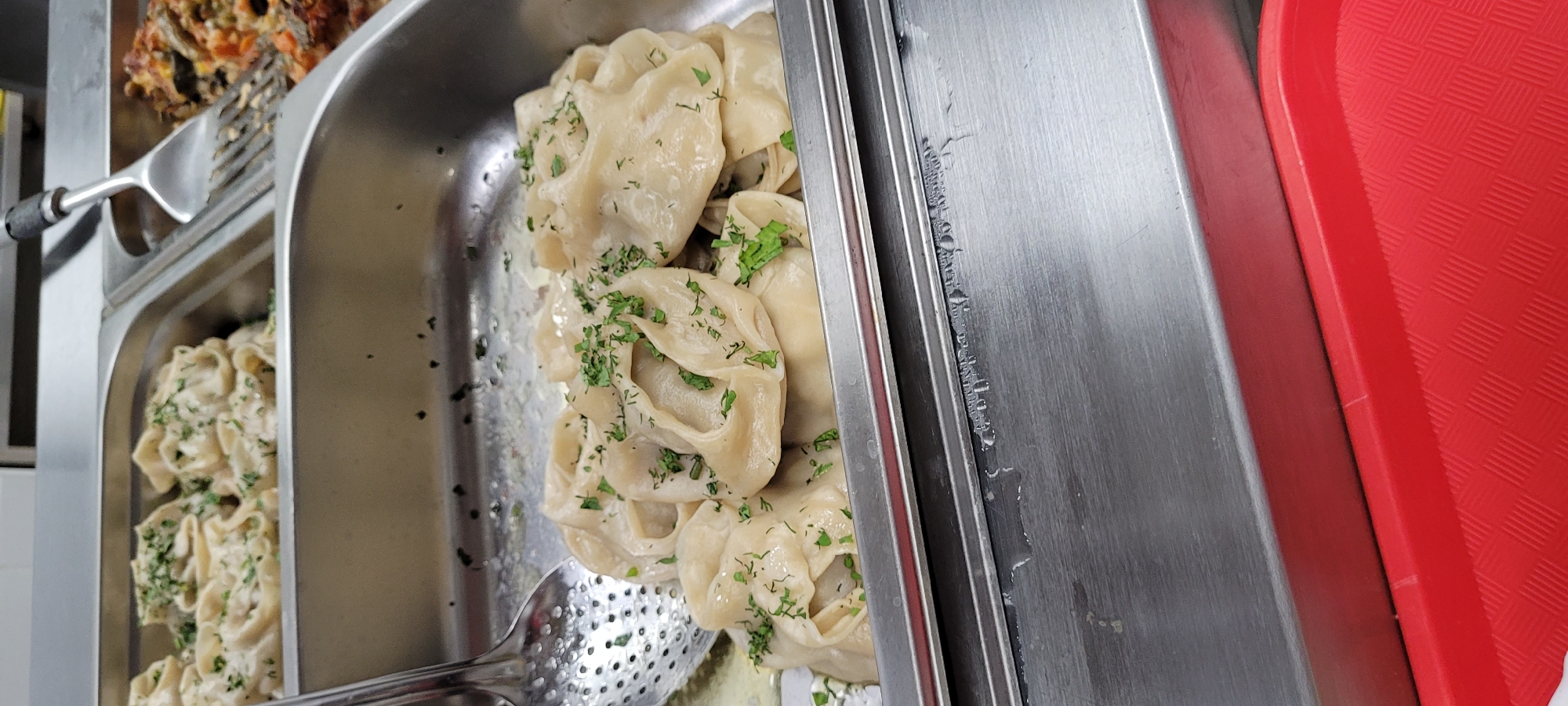 The image shows a stainless steel food warmer in an unknown location in the city of Unknown, Unknown. The warmer contains three food trays. The first tray contains a dish that appears to be pasta in a white sauce and garnished with herbs. The second tray contains a dish that appears to be manti, a type of dumpling, also garnished with herbs. A slotted spoon is partially visible in this tray. The third tray contains a dish that appears to be a baked vegetable dish. A stainless steel divider separates the food trays, and a red plastic tray is positioned next to the food warmer.