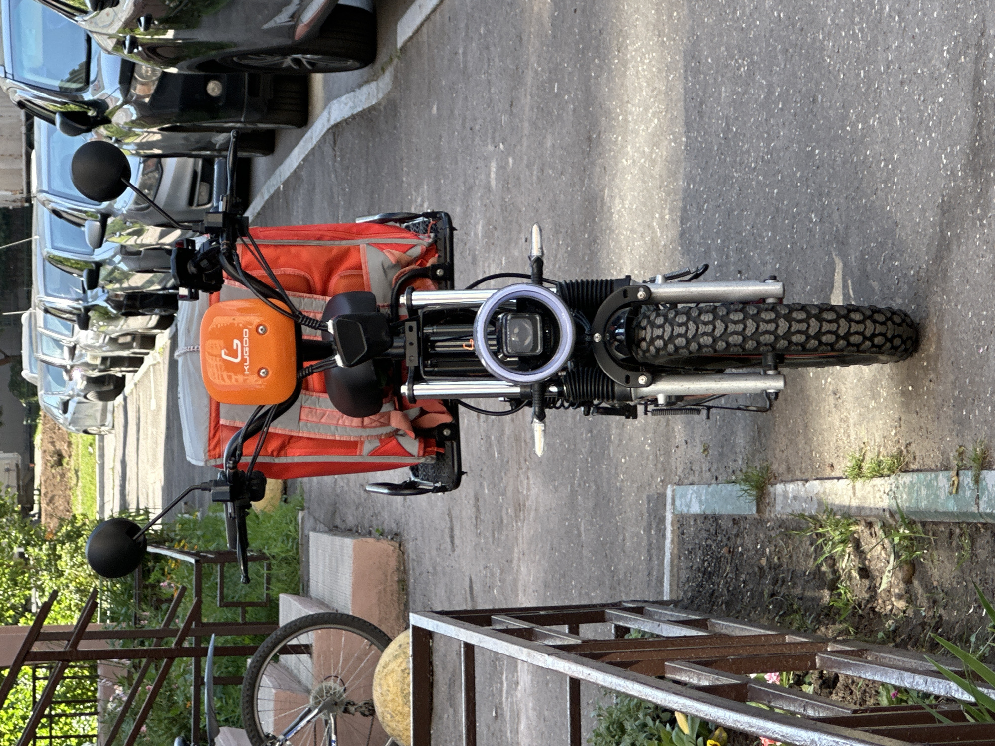A front-facing, eye-level, medium shot captures an electric scooter parked on an asphalt surface in what appears to be a residential courtyard in Podolsk, Russia. The scooter is orange and black, with a large red insulated delivery bag attached to the handlebars, suggesting its use for food or package delivery. A circular headlight with a white halo light is prominent on the front. The scooter's front tire, with a rugged tread pattern, is positioned centrally in the frame.
To the left of the scooter, a metal railing with a brown finish separates the scooter from a raised concrete structure and a patch of dirt with some green foliage. A bicycle wheel is partially visible behind the railing. In the background, a line of parked cars is visible, suggesting the presence of a road or parking area. The overall environment appears to be an outdoor urban setting under daylight.