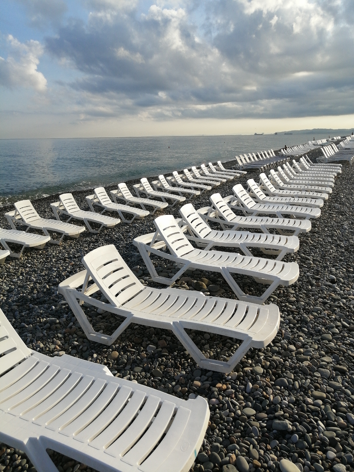 This is a wide-angle, eye-level shot of a nearly empty pebble beach in Batumi, Georgia, under a cloudy sky. The scene is dominated by rows of white plastic lounge chairs neatly arranged facing the calm, grey sea. The chairs are mostly unoccupied, with a few figures barely visible in the distance. The pebble shore is densely packed with small, grey, rounded stones. The water's edge is marked by a thin line of foam. In the far distance, ships can be seen on the horizon. The overall impression is one of quiet, post-season or pre-season emptiness on a seaside resort.