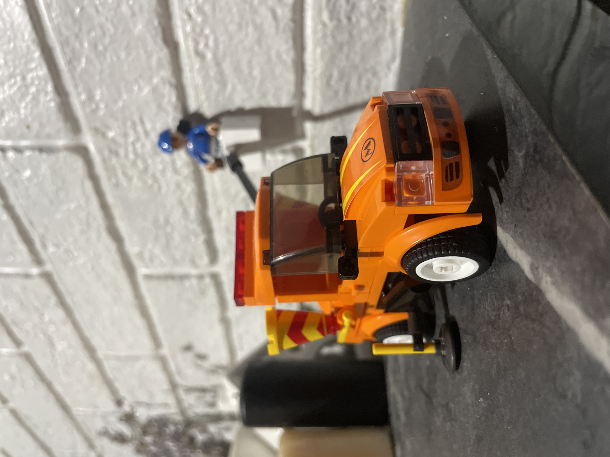 This media file shows a close-up, low-angle shot of an orange toy truck, possibly a construction or maintenance vehicle, resting on a dark, textured surface. The truck is positioned on its side, with its wheels visible. One of its rear wheels, with a white rim and black tire, is prominently displayed. The truck features a cabin with transparent orange windows, and on its side, there is an orange decal with a yellow lightning bolt symbol. Attached to the rear of the truck is a bright yellow and red striped cone or nozzle.
In the background, a white brick wall is visible, with some shadow patterns suggesting a light source. To the left of the truck, a small toy figure wearing a blue cap and a blue shirt is partially visible, standing near the brick wall. The overall scene appears to be indoors, possibly on a shelf or table, given the backdrop and the way the toy is positioned. No specific time of day or weather conditions can be determined. There is no visible text in the image. The location context provided is Baranovka, Russia, but this cannot be visually confirmed from the image itself.