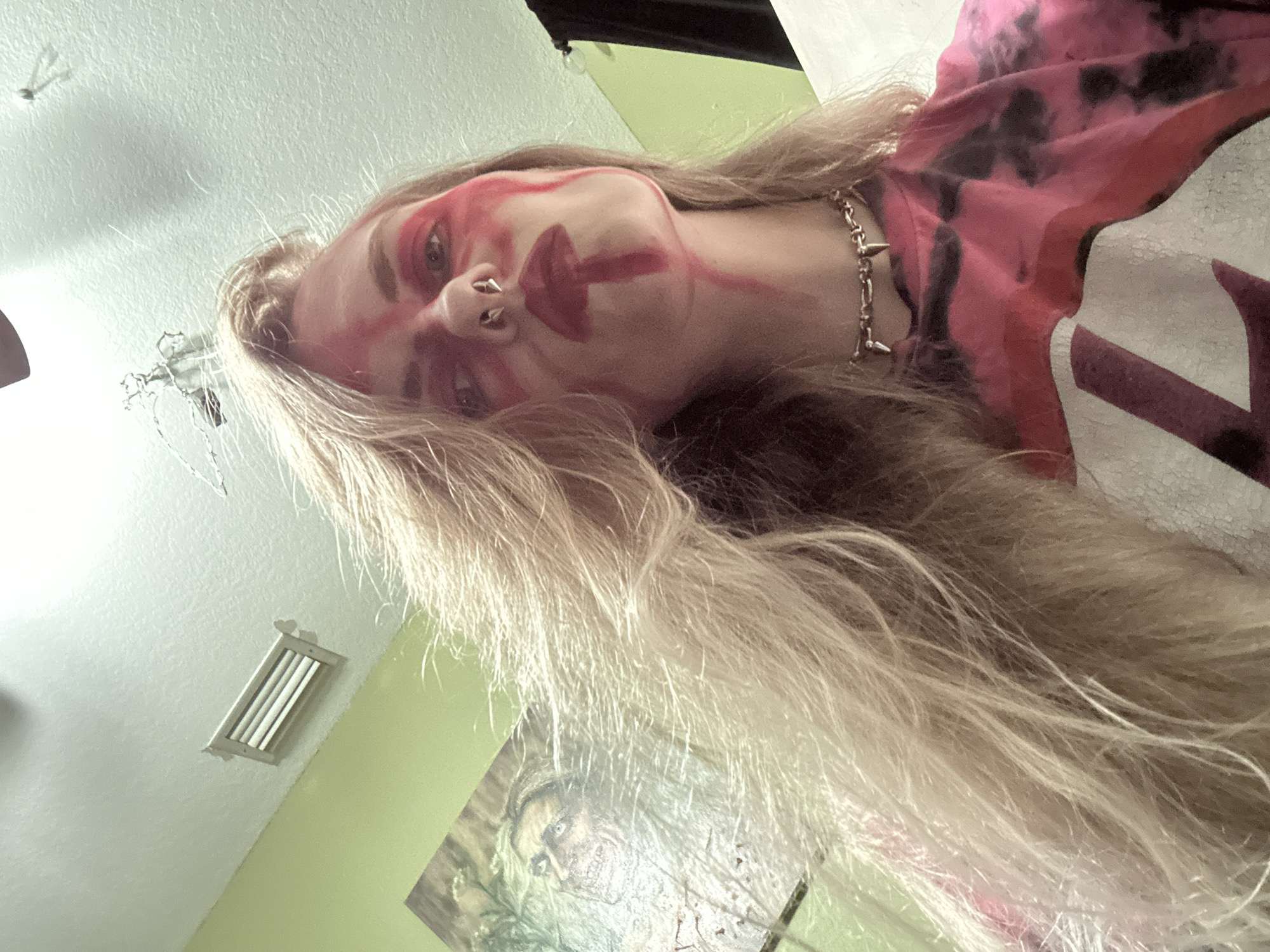 The image is a close-up, angled shot of a person with long blonde hair, possibly indoors. The person has red markings on their face, resembling tribal or abstract face paint. They have multiple piercings in their nose and are wearing a silver-colored, spiked choker necklace. A pink and black tie-dye shirt is partially visible. The background features a light green wall with a rectangular air vent and a poster or artwork depicting a zombie-like figure. The location is within the city of Cape Coral, United States of America.