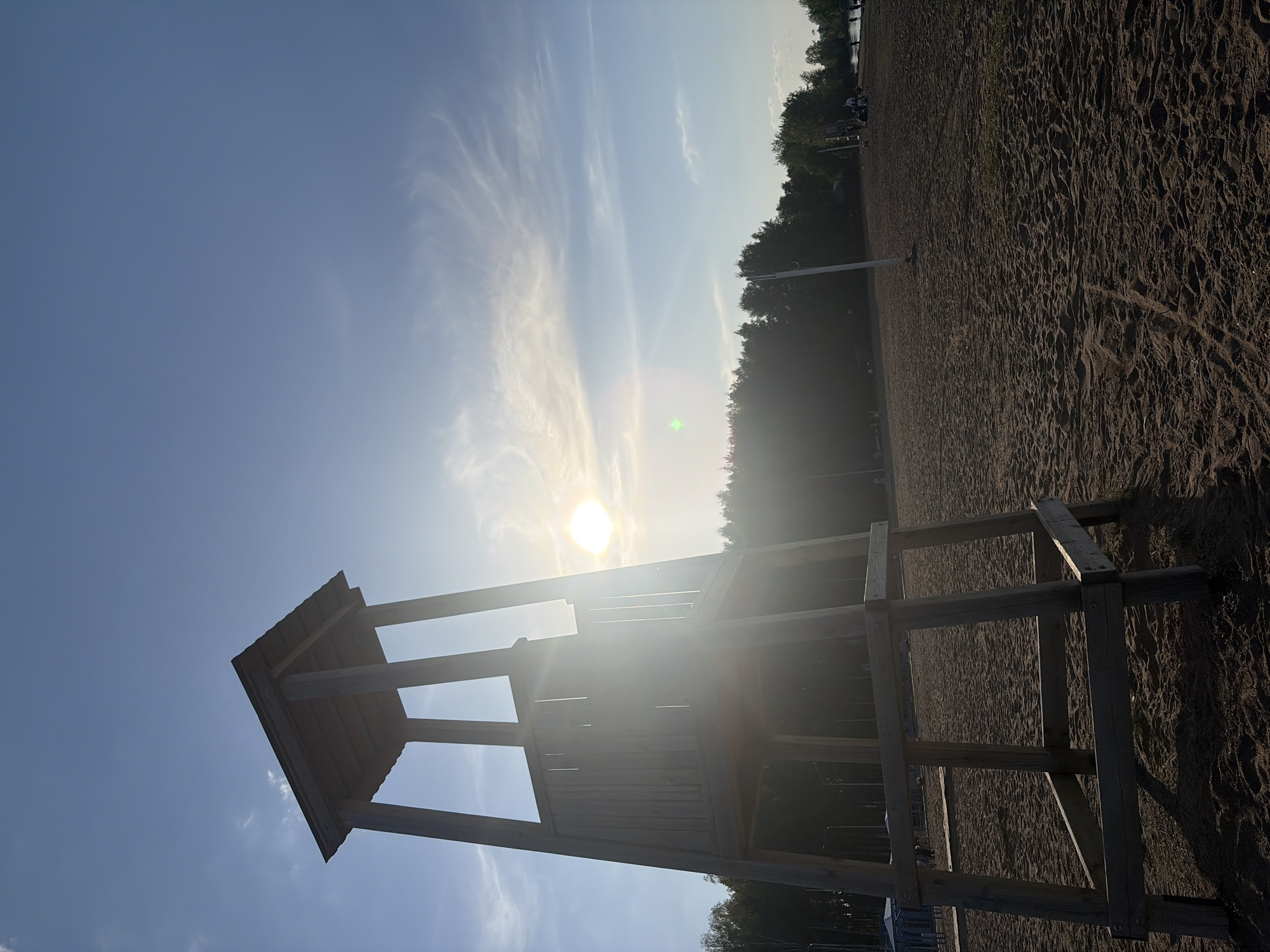 The image shows a lifeguard stand on a sandy beach with the sun setting or rising in the background. The sky is clear with some wispy clouds. Trees are visible along the horizon. The lifeguard stand appears to be made of wood. The image was taken in Chaikovsky, Russia.