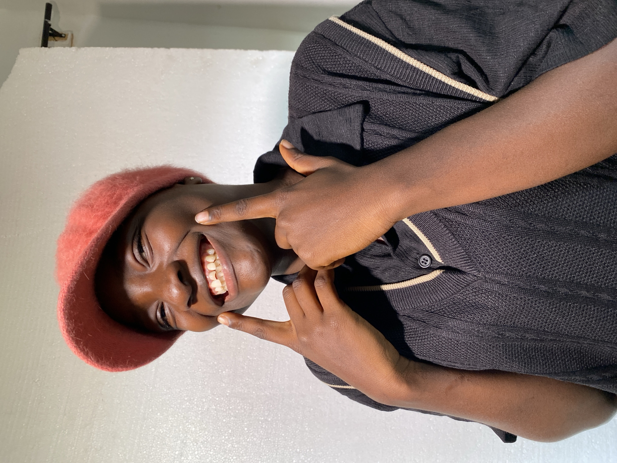 A person wearing a pink beret and a dark collared shirt with cream stripes is shown. They are smiling widely, showing their teeth, and have their hands on their face. The person's fingers are pointing towards their cheeks, and they are making a "rock on" gesture with their other hand. The background is a plain, textured white surface. The image is well-lit, suggesting it was taken indoors or with studio lighting. The person's skin tone and the style of clothing suggest a warm climate. The person appears to be happy and playful. No text is visible in the image. The location is identified as Jos, Nigeria.