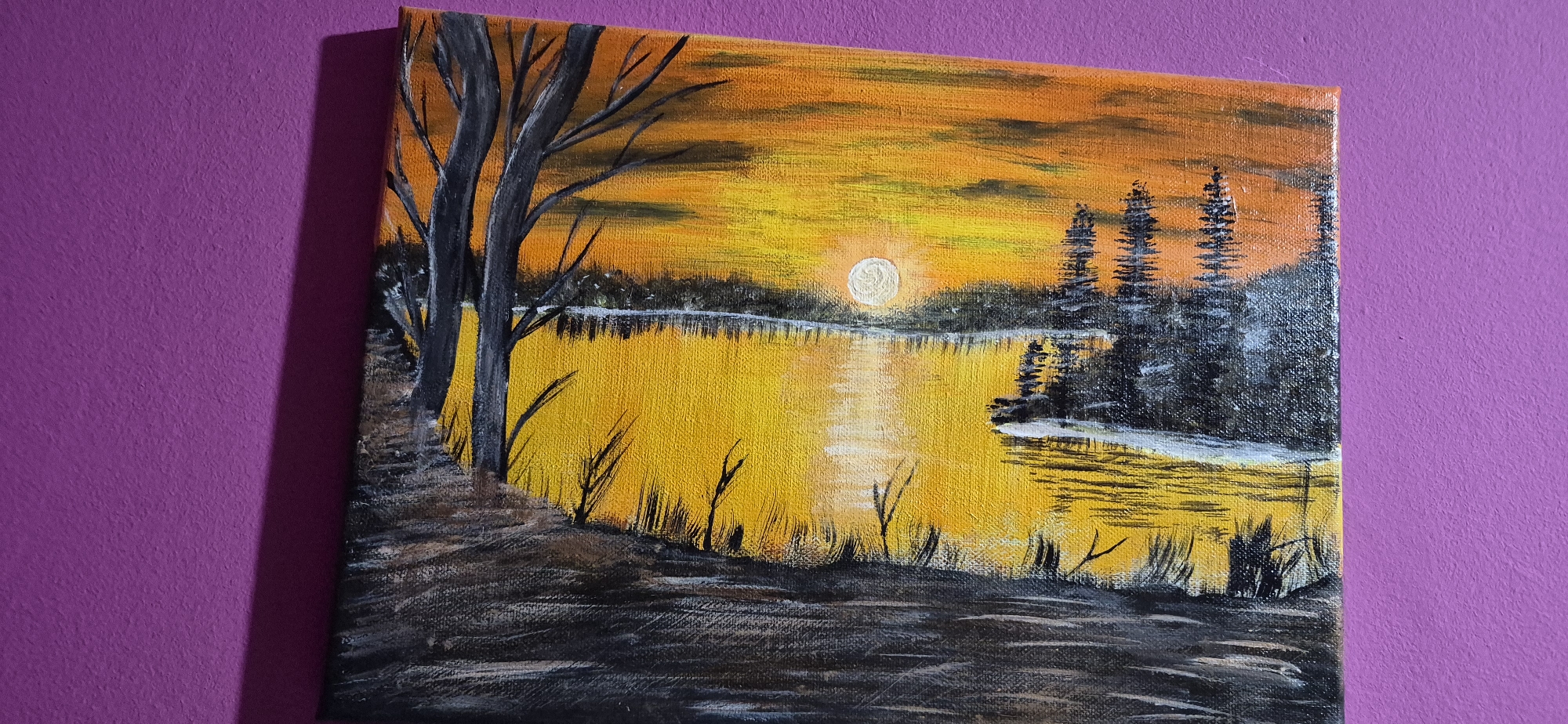 The image shows a painting of a sunset over a body of water. The painting is framed and hanging on a purple wall. The painting depicts a landscape with a lake or river reflecting the vibrant orange and yellow hues of the setting sun. Silhouetted trees line the far bank, and there are trees and foliage in the foreground. The overall impression is one of a serene, natural scene at dusk. There is no indication in the image or the provided context that this painting is located in Lom, Bulgaria, or any other specific city. The image only shows the painting itself and the wall it is hanging on.