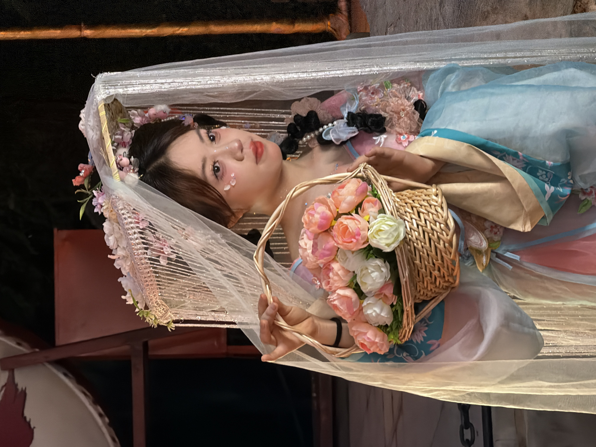 A young woman is depicted lying down, adorned with a floral headdress and delicate makeup that includes small, shimmering embellishments on her cheeks. She is wearing a flowing, layered gown in pastel shades of blue and pink, possibly a traditional Chinese garment or a costume inspired by one. A small wicker basket filled with pink and white artificial flowers rests in her hands. She is positioned within a structure draped with translucent, sparkly fabric, giving the impression of a delicate canopy or a theatrical setting. The lighting suggests an indoor environment, possibly a studio or a themed event. The overall mood is serene and ethereal, with the woman's expression being soft and contemplative. The background is dark and out of focus, highlighting the subject. The image appears to be taken in Yuzhong District, China, though no specific landmarks are visible.
