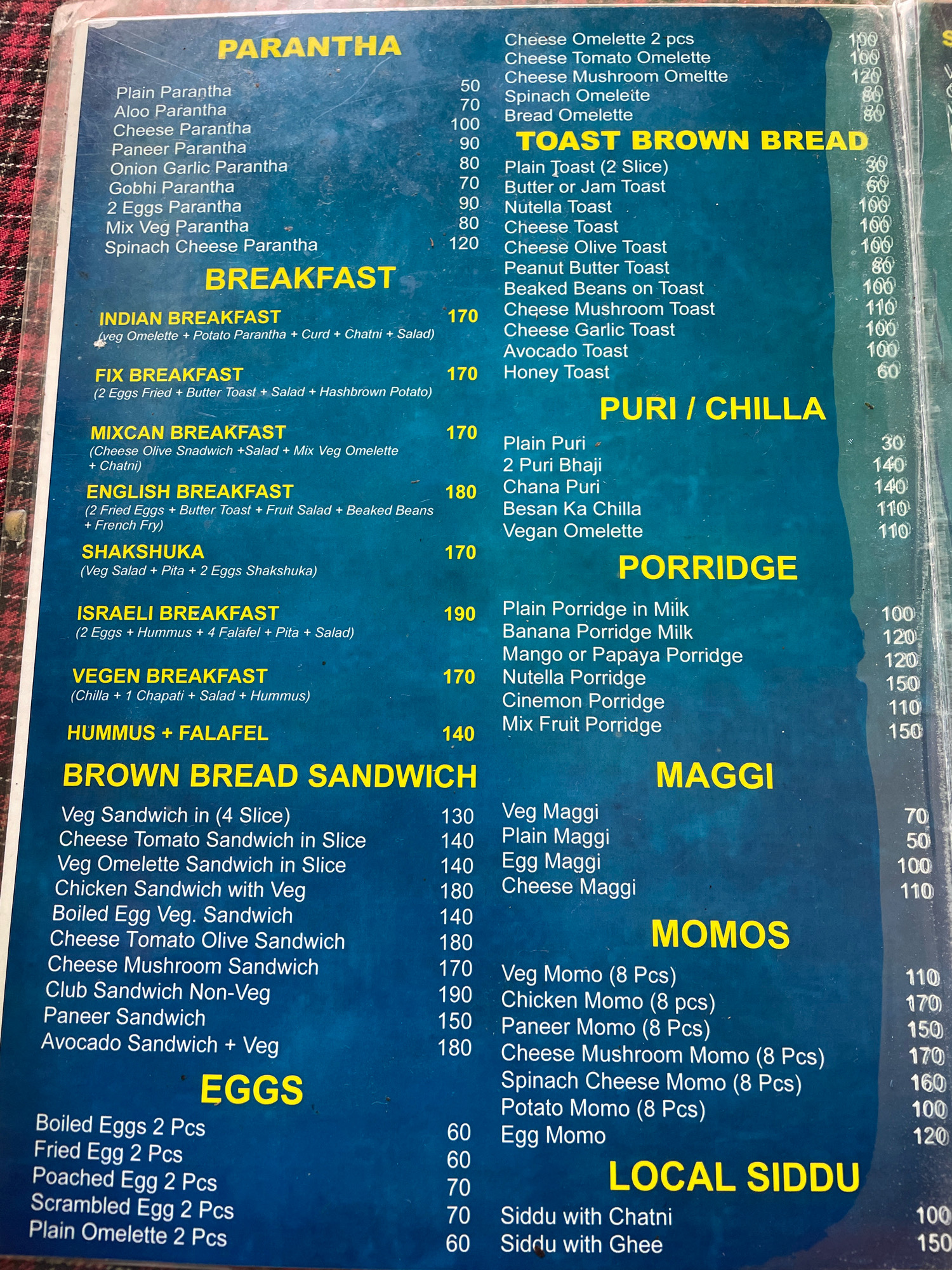 The image shows a menu from a restaurant in Vashisht, India. The menu is printed on a blue background and lists various food items, including paranthas, toast brown bread, puri/chilla, porridge, brown bread sandwiches, eggs, maggi, momos, and local siddu. Each item is accompanied by its price. The lighting is even, and the menu is viewed from a slightly overhead angle. There are no people or other objects visible in the image, other than the menu itself and the surface it is resting on, which appears to be a textured, possibly wooden, surface. The image is a close-up of the menu, suggesting it was taken to document the offerings and prices.