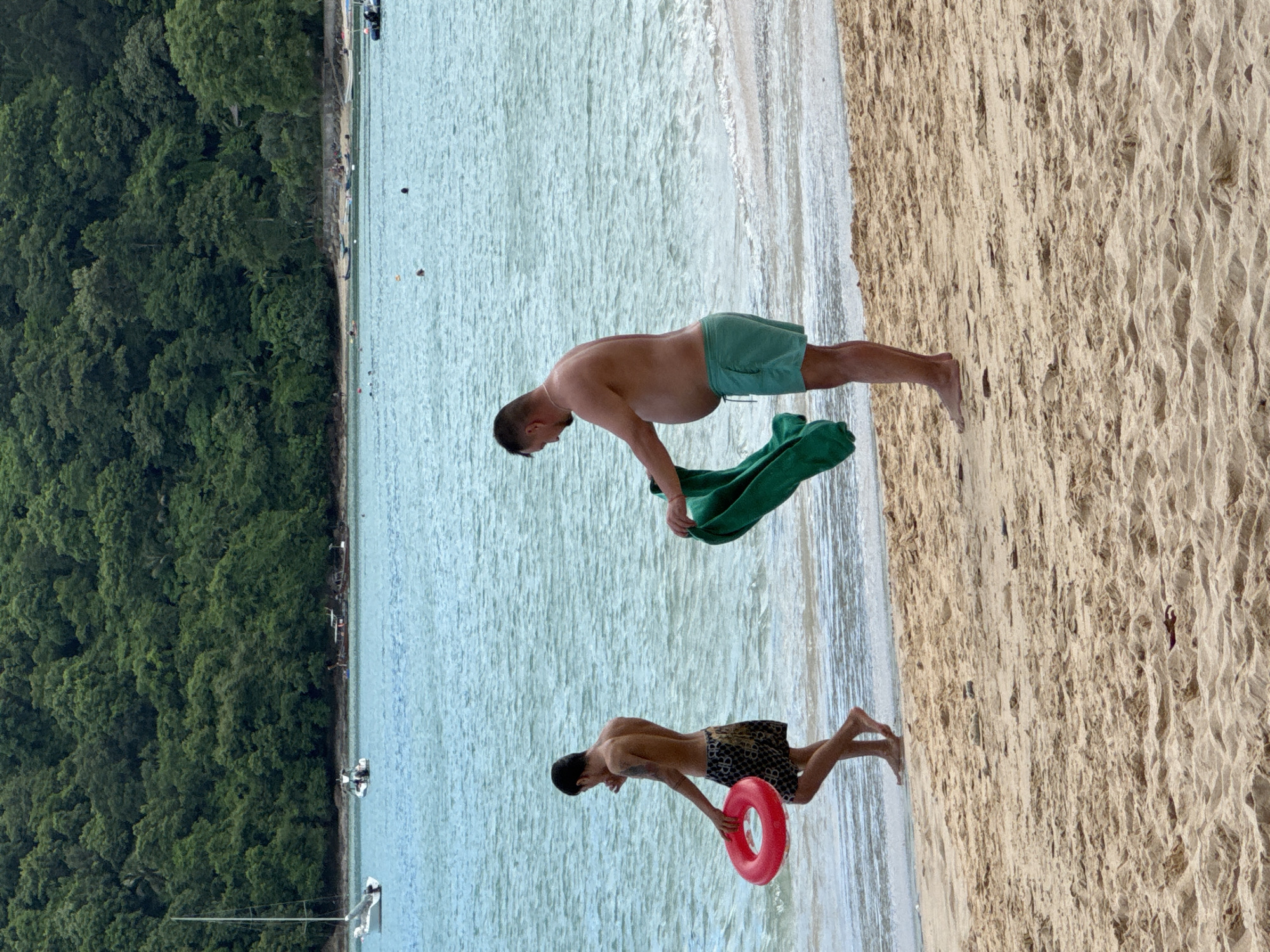 Two men are on a sandy beach in Wichit, Thailand. The man on the left, facing away from the camera, is wearing patterned swim trunks and holding a red inner tube. The man on the right, facing left, is wearing green swim trunks and holding a green towel. He has a large belly. The ocean is behind them, with gentle waves lapping at the shore. In the distance, a lush green hillside covered in trees rises from the water. Several boats are visible on the calm, blue water. The sand on the beach is light brown and textured.