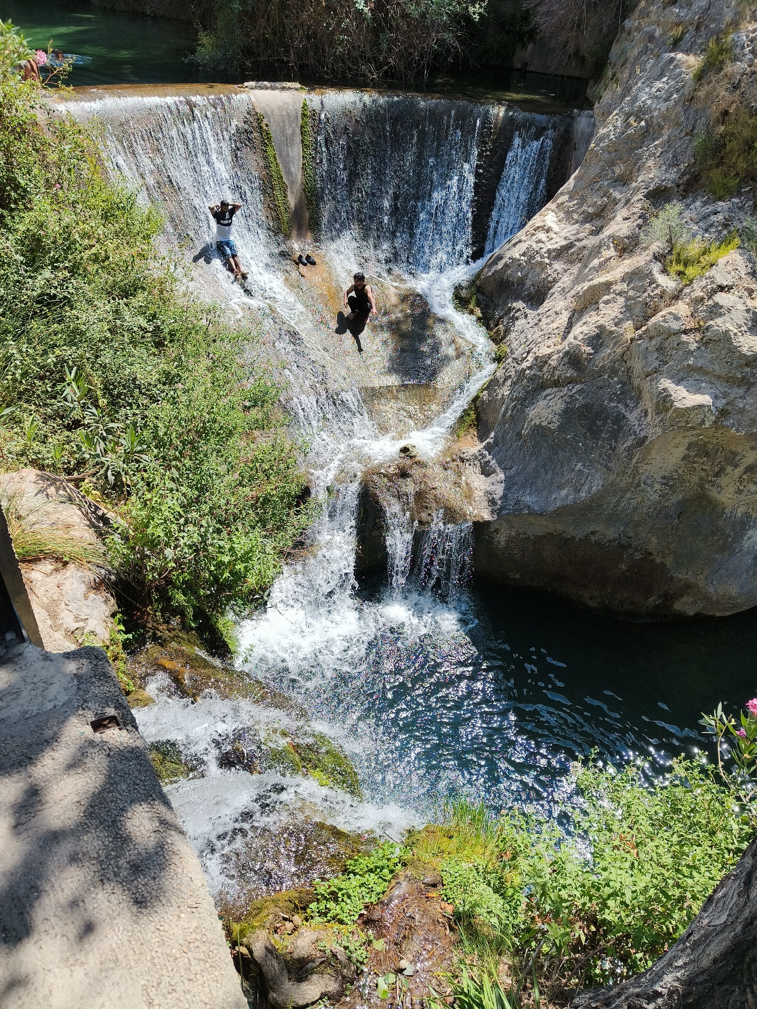 The image shows a natural waterfall in a rocky, vegetated area. There are several people present. In the foreground, a person is sliding down the waterfall. Further up, another person is seated at the top of the waterfall. In the background, two people are in the water. The environment is lush with green vegetation surrounding the water. This natural scene is located in the Pou Clar of Ontinyent, Spain, a natural water park with several natural pools and cascades formed by the Clariano River.