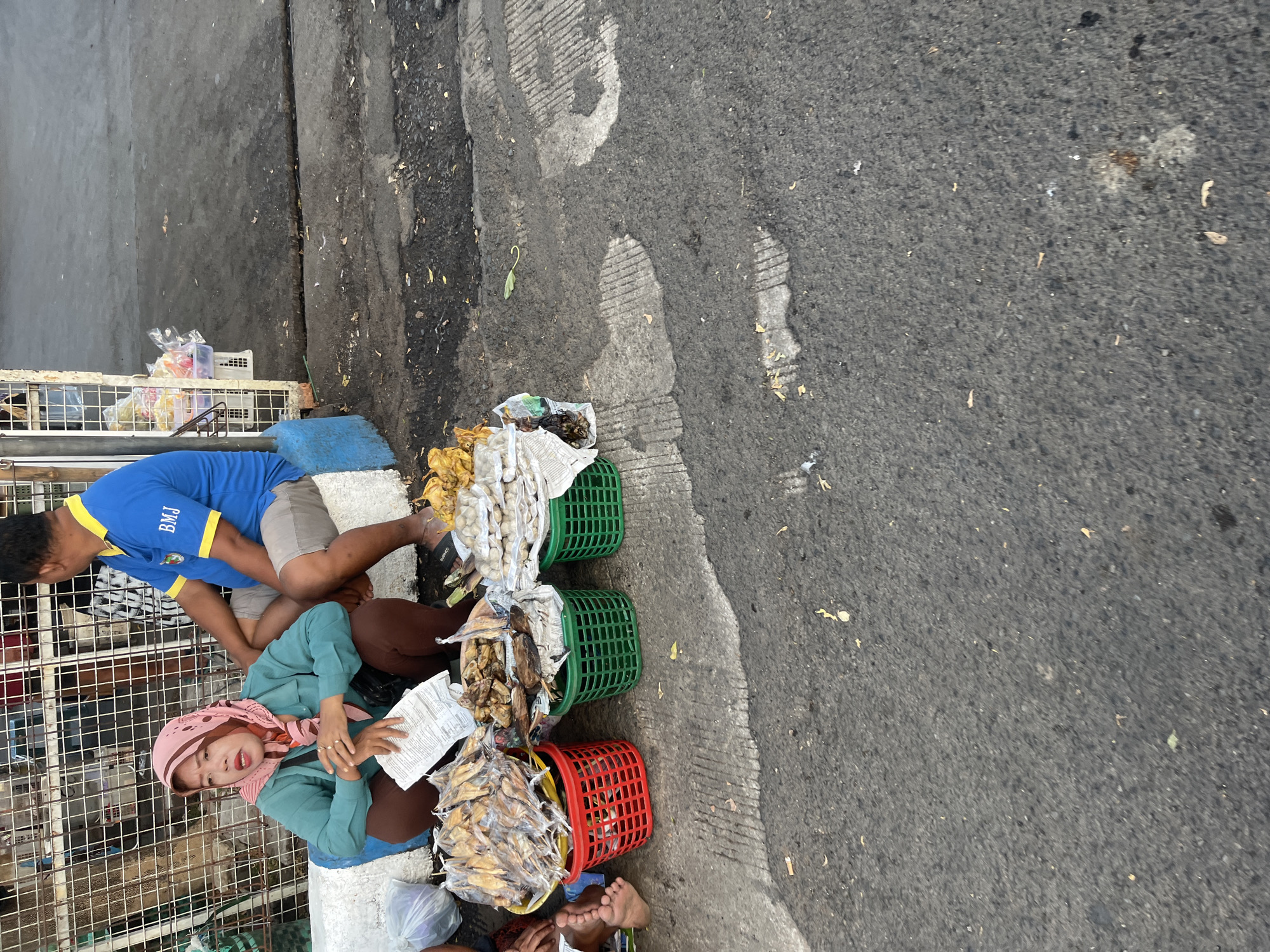 The image depicts a street scene in Pelang, Indonesia. A woman wearing a pink hijab and a turquoise shirt sits on the ground in front of a display of dried fish and other food items packaged in plastic bags and baskets. To her right, a man in a blue t-shirt with yellow trim sits next to her. The food is arranged in red, green, and yellow baskets. In the background, a white fence with wire mesh encloses a stall. The ground is asphalt, with some debris and cracks visible. The lighting suggests it is daytime, possibly late afternoon given the shadows.