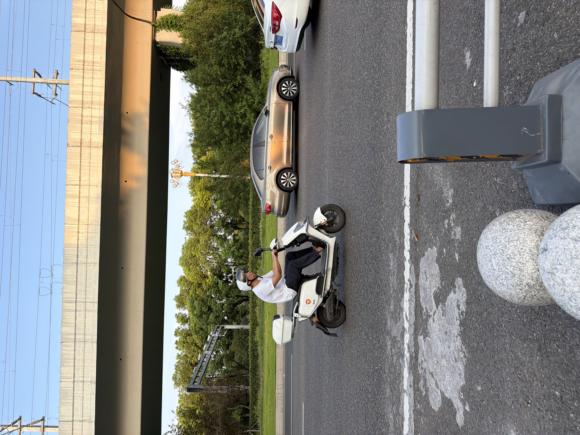 In the city of Jiangjin, China, a man is riding a white electric scooter on a paved road. He is wearing a white t-shirt, dark pants, and a white helmet. The scooter has a white storage box on the back. To his right, a gold sedan is driving in the same direction. Further to the right, the rear of a white car is visible. The road is lined with trees and greenery, and an elevated concrete structure with overhead wires is visible in the background. The scene appears to be a street in an urban or suburban area, possibly during the late afternoon, given the warm lighting.