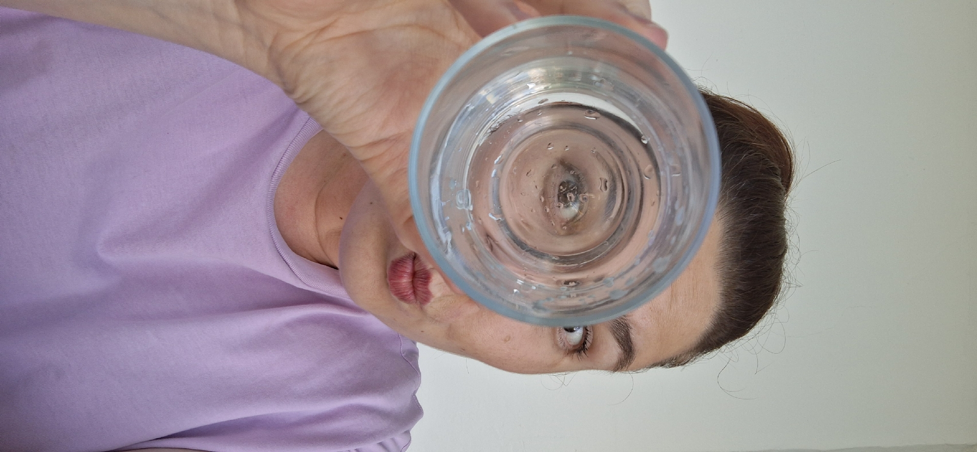 A woman is holding a glass of clear liquid up to the camera, capturing her reflection in the liquid. She's wearing a light purple t-shirt and has her hair pulled back. The background appears to be a plain, light-colored wall. It is impossible to verify that the image was taken in Chaikovsky, Russia.