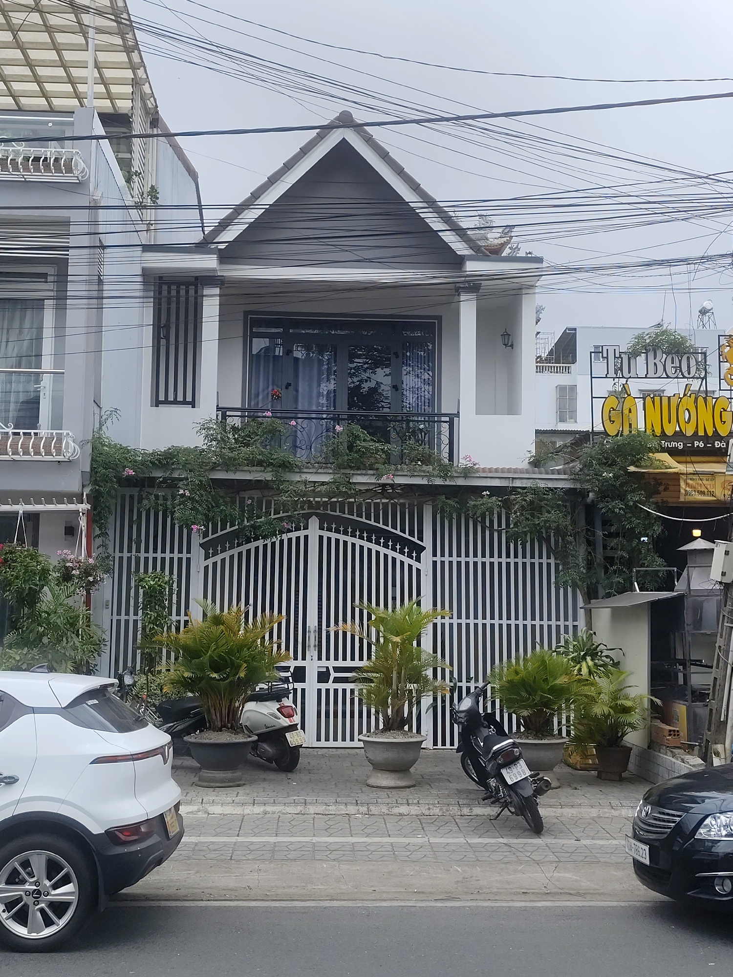 In Da Lat, Vietnam, a white SUV is parked on a sidewalk in front of a two-story building. The building has a white facade, a dark-tiled roof, and a balcony on the second floor with black railings. A white metal gate in front of the building is partially obscured by lush green plants that climb along the facade. Two motorcycles are parked behind the gate. A neon sign in yellow and white, reading "Tu Beo Ga Nuong" which translates to "Tu Beo Grilled Chicken", is visible on the right side of the image, indicating a food establishment. The street is paved with grey tiles, and power lines crisscross the sky. The weather appears overcast.