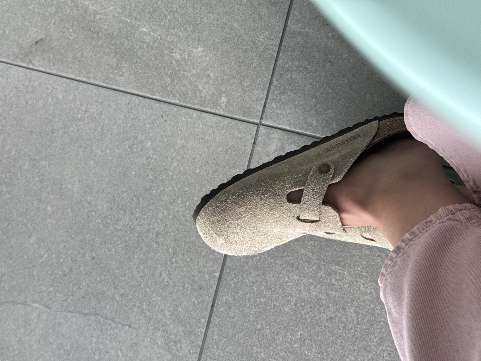 The image presents a close-up, angled view of a person's left foot wearing a light beige suede Birkenstock shoe. The shoe is a closed-toe style, possibly a clog or a Mary Jane, featuring a strap over the instep and a dark, textured sole. The brand name "BIRKENSTOCK" is clearly visible in black lettering on the inner sole near the arch of the foot, accompanied by a small circular logo.
The person's leg is partially visible, clad in a light pink garment, likely a pant leg, with a visible ribbed or corduroy texture and a cuffed hem. The foot rests on a light gray tiled floor, composed of rectangular tiles with distinct dark grout lines forming a grid. The tiles exhibit a subtle speckled texture. In the upper right portion of the frame, a smooth, light blue or light green object creates a bright reflection, partially obscuring the background. This object could be part of a wall or a piece of furniture.
The scene depicts an indoor setting, likely during the daytime, given the bright and consistent illumination. There are no overt activities taking place, just the static display of the foot and shoe. The image was captured in San Bernardino Tlaxcalancingo, Mexico.