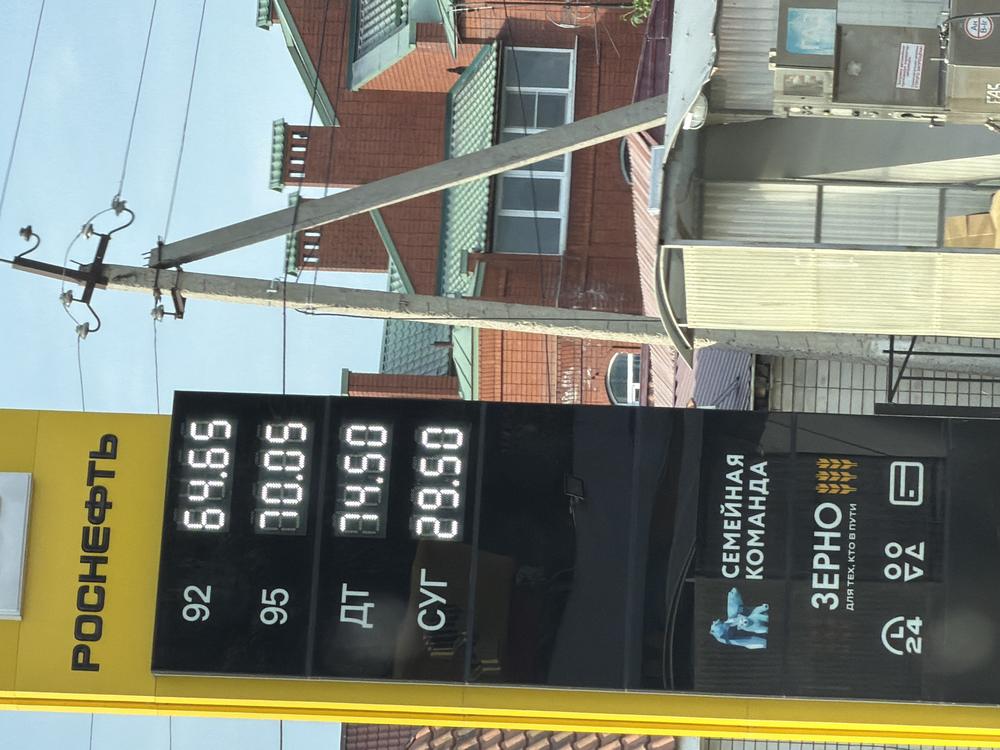 The image shows a close-up of a yellow and black electronic price board at a Rosneft gas station in Ust-Dzheguta, Russia. The board displays fuel prices: 92 octane gasoline at 64.65 rubles, 95 octane gasoline at 70.05 rubles, diesel fuel (DT) at 74.50 rubles, and a fuel identified by the Cyrillic abbreviation "CYГ" at 29.50 rubles. Below the fuel prices, there are several logos and text. One logo depicts polar bears with the text "СЕМЕЙНАЯ КОМАНДА" (Family Team). Another section has the word "ЗЕРНО" (Grain) with the tagline "ДЛЯ ТЕХ, КТО В ПУТИ" (For those who are on the road), accompanied by an illustration of wheat stalks. Further down are icons representing 24-hour service, a payment method (possibly indicating card acceptance), and a credit card. In the background, a utility pole with insulators and wires stands against a bright sky. Behind the pole, there are red brick buildings with green tiled roofs, characteristic of residential structures in the region. Part of a corrugated metal structure is also visible in the foreground. The overall scene captures a typical view of a gas station and its surroundings in a Russian town.