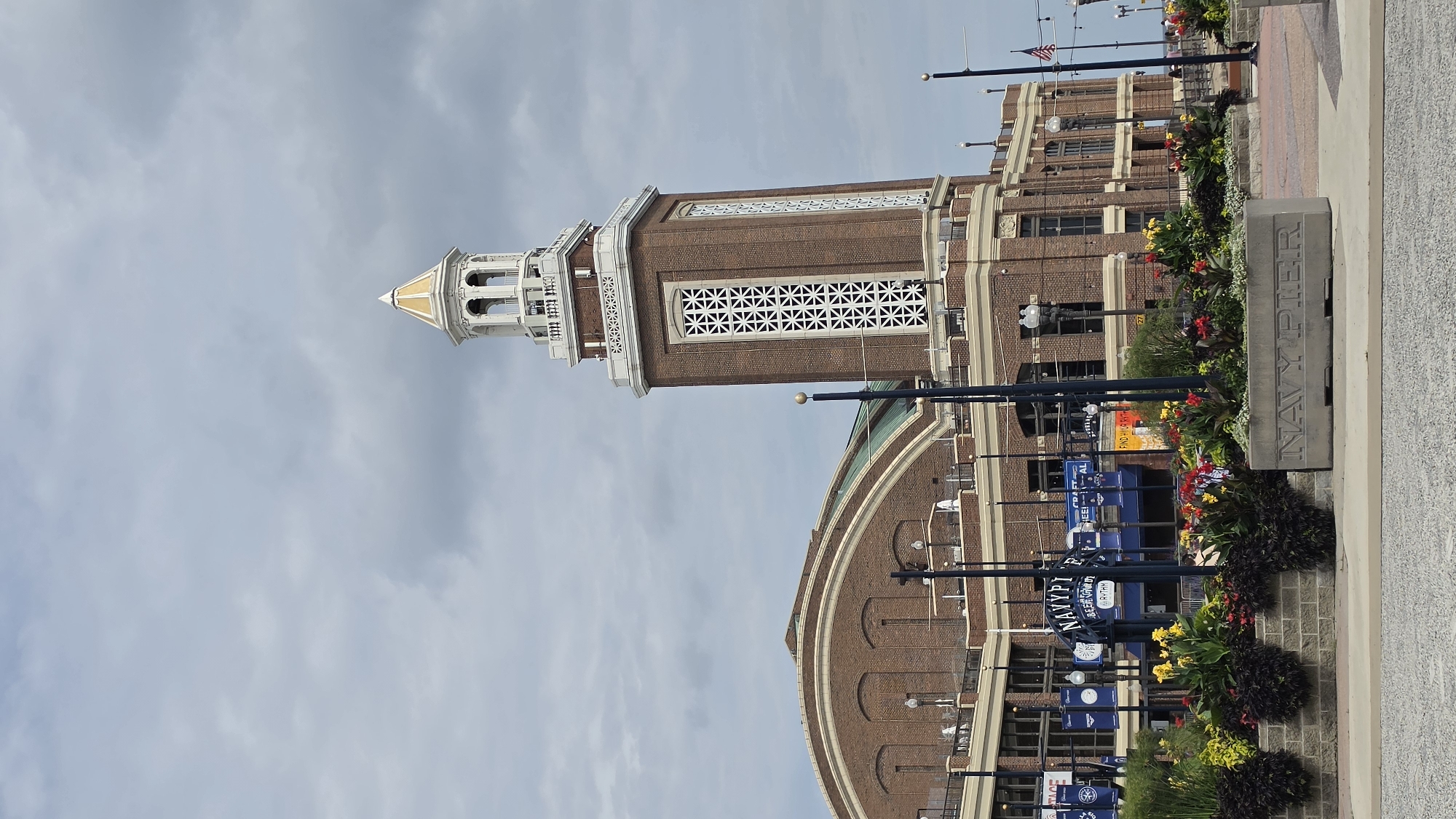 The image captures a view of Navy Pier in Chicago, Illinois, under a cloudy sky. In the foreground, a concrete planter box with "NAVY PIER" etched into it sits on a paved area. Beyond the planter, a vibrant flowerbed bursts with red, yellow, and dark purple blooms. Behind the flowers, a building complex made of red brick is visible. The complex features a prominent tower with a white decorative top and a gold spire. The tower has a tall, narrow design with patterned windows. To the left of the tower, a large, arched section of the building with a green roof is visible. Numerous lampposts are scattered throughout the scene. Several flags, including an American flag, are also present. The overall environment appears to be an outdoor public space.