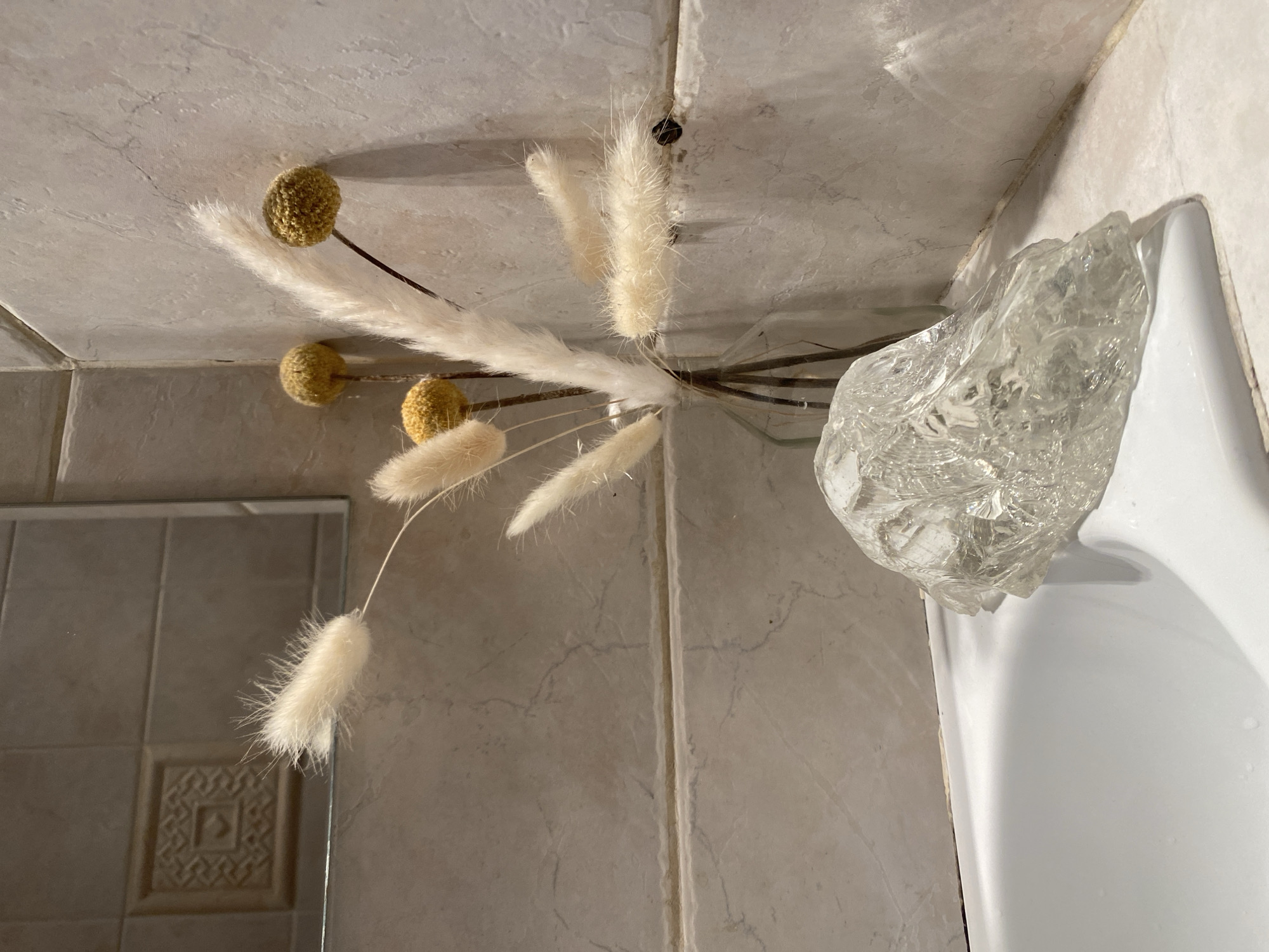 The image shows a close-up view of a bathroom corner. The walls and backsplash are tiled in a light beige marble pattern. A bouquet of dried flowers with feathery plumes and spherical yellow heads is arranged in a clear, textured glass vase. The vase sits on the edge of a white porcelain sink or bathtub. A mirror is visible in the lower left corner. The location is not specified and could not be determined from the image.