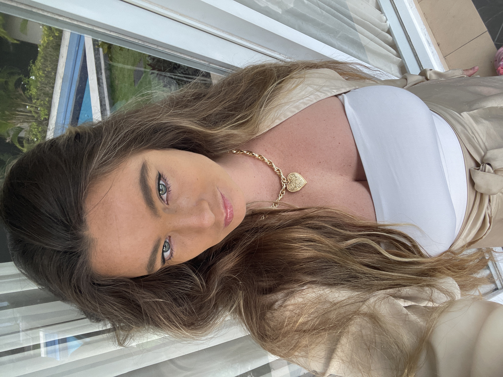 A woman with long, wavy brown hair is taking a selfie on a balcony with a pool visible in the background. She is wearing a white tube top, a gold shimmering jacket, and a gold heart-shaped pendant necklace. The setting appears to be a tropical location, possibly a resort, given the presence of the pool and lush greenery. The woman is looking directly at the camera with a slight smile.
