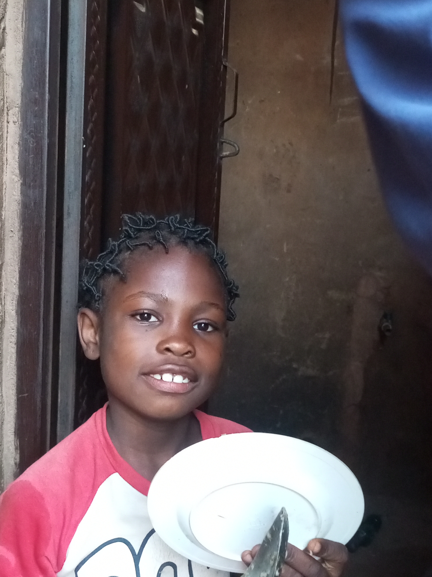 A young girl with dark skin and braided hair smiles at the camera, holding a white plate and a knife. She is wearing a red and white t-shirt. The background appears to be an outdoor setting with a wooden doorframe and a textured wall. The lighting suggests it is daytime. The image was taken in Tempure, Nigeria.