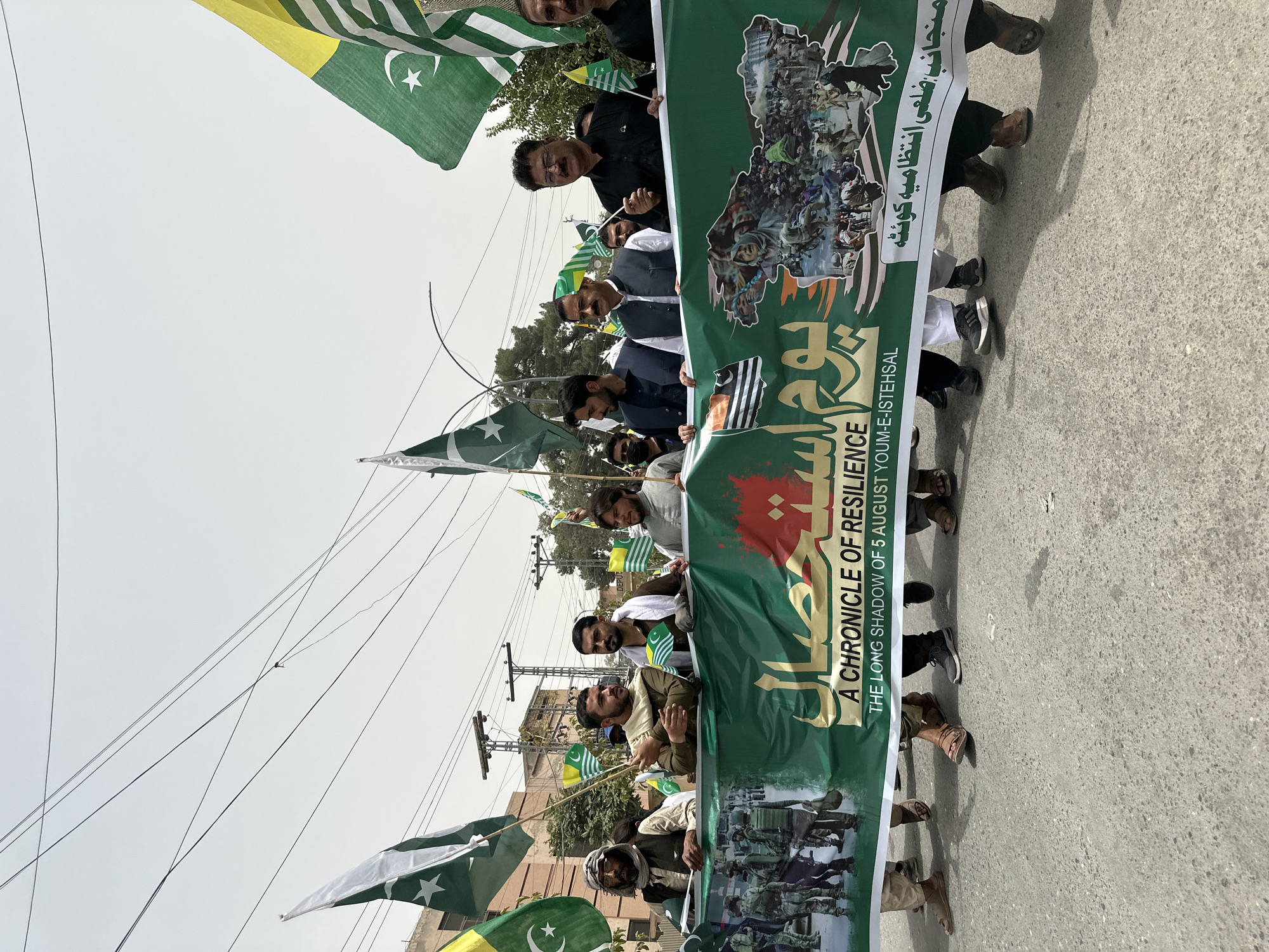 In Quetta City Tehsil, Pakistan, a group of men are participating in a procession, holding a large green banner. The banner features images of people and soldiers, along with text in both English and Urdu. The English text reads "A CHRONICLE OF RESILIENCE THE LONG SHADOW OF 5 AUGUST YOUM-E-ISTEHSAL". Several flags are also visible, including the flag of Pakistan and what appears to be the flag of Azad Kashmir. The men are dressed in a variety of clothing, including traditional shalwar kameez, vests, and head coverings. The procession is taking place on a paved street, with buildings and utility poles visible in the background. The sky is overcast.