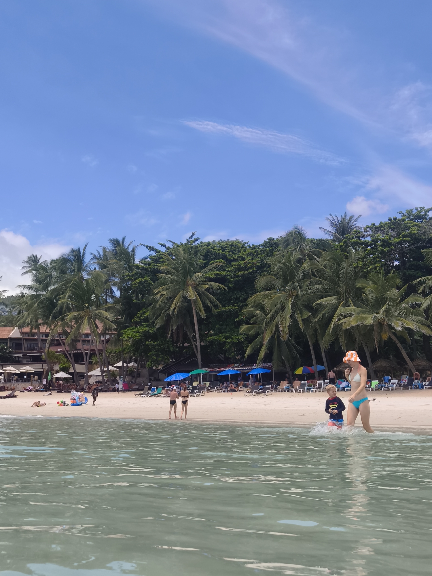 A bright, sunny day at a tropical beach. The water in the foreground is clear and shallow, with gentle ripples reflecting the light. In the middle ground, a woman in a white bikini and an orange-striped sun hat walks through the water, followed by a young boy wearing a blue rash guard with a yellow symbol and red shorts. They appear to be enjoying a leisurely stroll at the edge of the sea.
The sandy beach behind them is populated with numerous beach chairs and colorful umbrellas, indicating a popular tourist destination. A line of lush palm trees and dense green foliage forms a backdrop to the beach, with a glimpse of buildings nestled amongst the greenery, suggesting resort accommodations. Several people are visible on the beach, some relaxing on loungers, others strolling. In the distance, the sky is a vibrant blue with scattered white clouds, indicative of fair weather. The overall scene conveys a sense of relaxation and vacation enjoyment. The location is identified as Ko Samui District, Thailand, which is consistent with the tropical scenery. No visible text is present in the image.