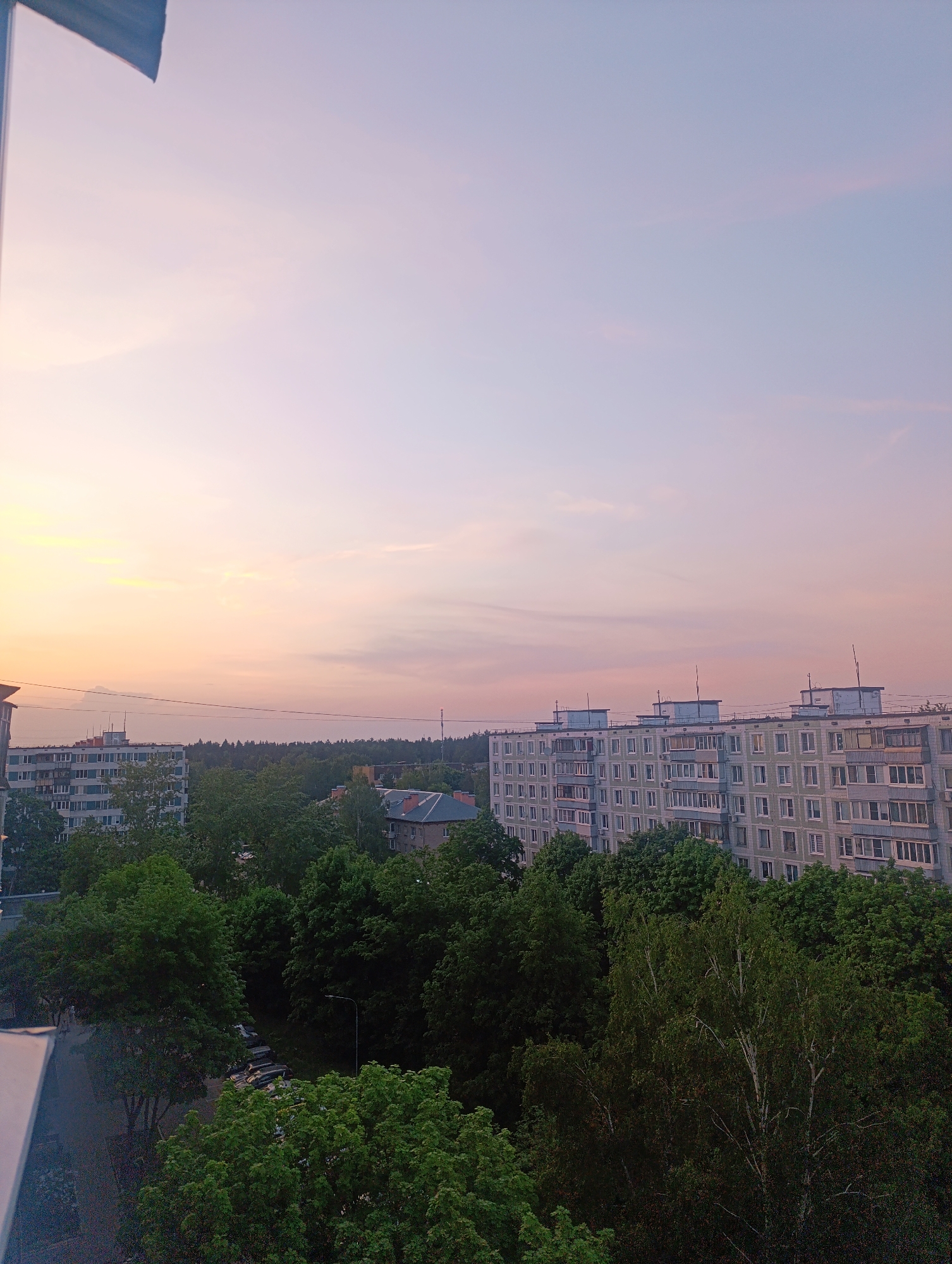 The image depicts a cityscape under a pastel-colored sky transitioning from sunset hues near the horizon to lighter purples and blues higher up. In the foreground, dense clusters of green trees fill the lower portion of the frame, obscuring some of the urban landscape. Behind the trees, there are apartment buildings, characterized by their rectangular shapes and repeated patterns of windows. The building on the right is taller, with multiple stories and a light-colored facade, while another similar building is visible further to the left. In the middle ground, a denser patch of trees forms a treeline against the backdrop of the sky. A street lined with parked cars can be seen at the bottom left. A small portion of a window frame or balcony is visible on the left edge of the image. Overall, the scene presents a tranquil urban environment at dusk. The location is unknown.