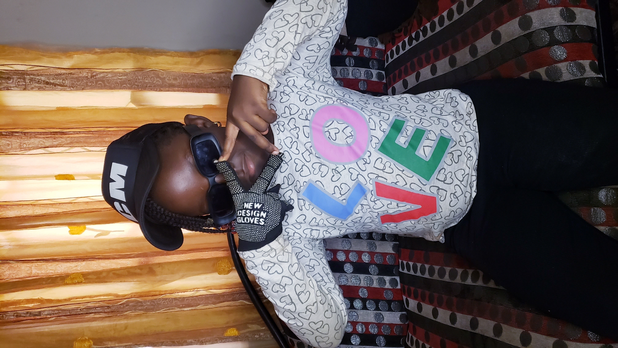 A dark-skinned individual, seemingly female, lies on a striped sofa under a gold-colored patterned drape or curtain. The person is wearing a black cap with white lettering (partially obscured), large sunglasses, a patterned long-sleeved shirt with the word "LOVE" in large colorful letters, and black pants. They are making a peace sign with a gloved hand; the glove reads "NEW DESIGN GLOVES". The location is within Narayi, Nigeria. The person is posing.