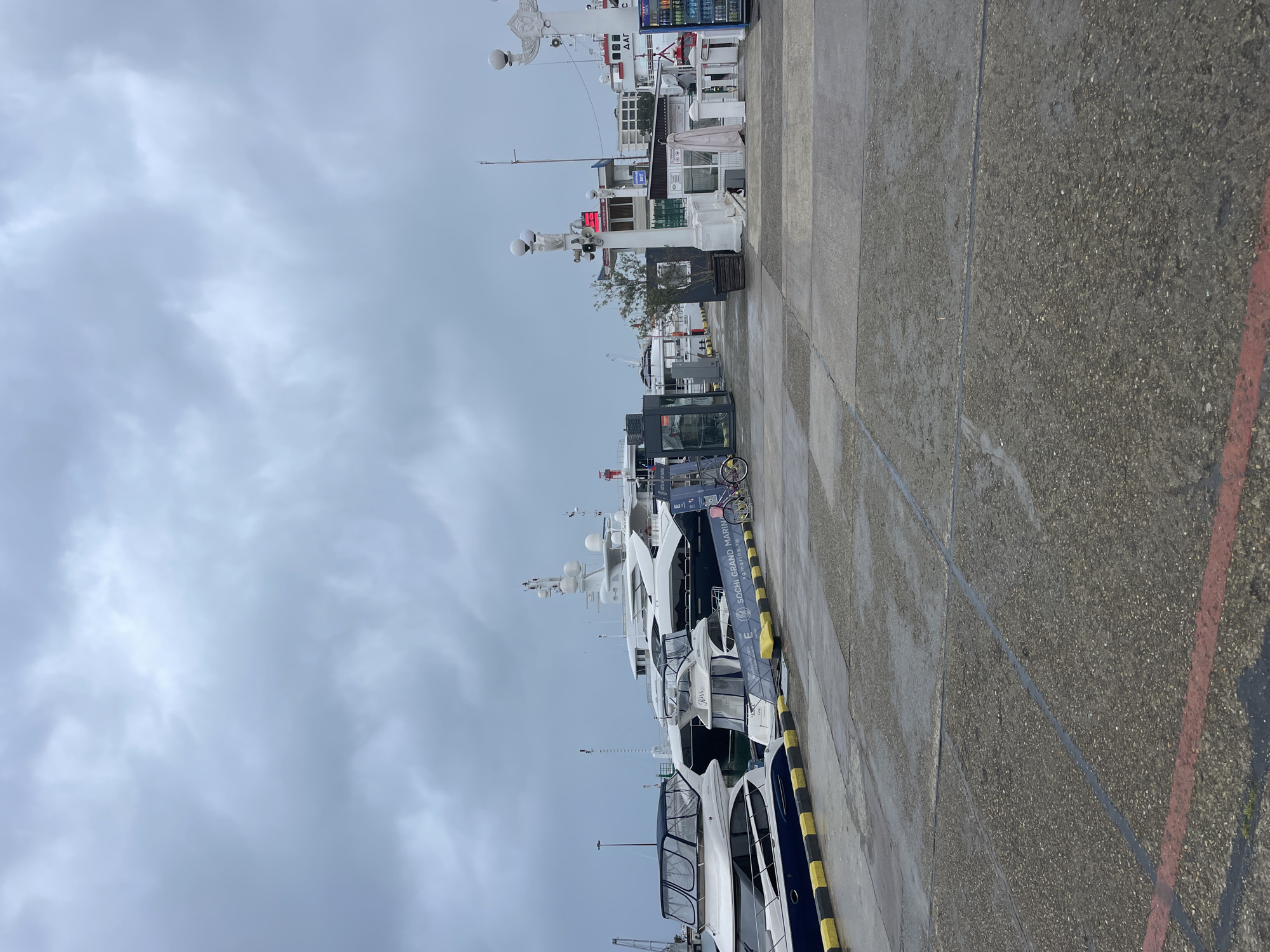 The image shows a row of yachts docked at a marina in Sochi, Russia. The sky is overcast. A concrete walkway runs along the side of the boats, marked with yellow and black stripes. A bicycle is parked near one of the yachts. There are no people visible in the shot.