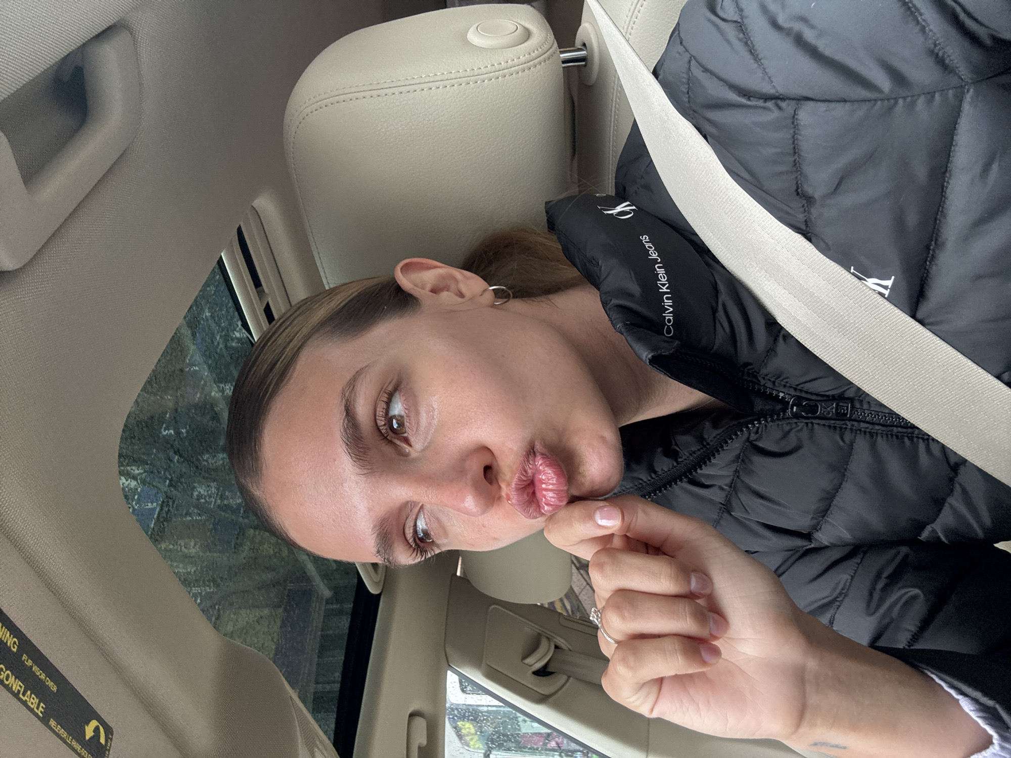A young woman is captured in a close-up, selfie-style photograph, situated inside a car. Her head is tilted, and she is making a "pouty" expression with her lips, looking towards the camera with her brown eyes. Her light brown hair is pulled back, revealing a silver hoop earring in her left ear. She wears a black puffer jacket with the "Calvin Klein Jeans CK" logo visible on the sleeve, and a light-colored seatbelt crosses her chest. Her right hand, adorned with a ring on one finger and light-colored nail polish, is gently placed near her chin and mouth.
The car's interior is light-colored, featuring beige leather seats and fabric ceiling. A headrest and a grab handle are visible in the background. Through the car window, blurred greenery and what appear to be raindrops suggest an outdoor, possibly overcast, and wet environment. A sun visor in the car displays warning text, partially visible as "WARNING FLIP VISOR OVER" and "GONFLABLE RELEVER LE PARE-SOLEIL". The overall impression is a casual moment captured during a drive.