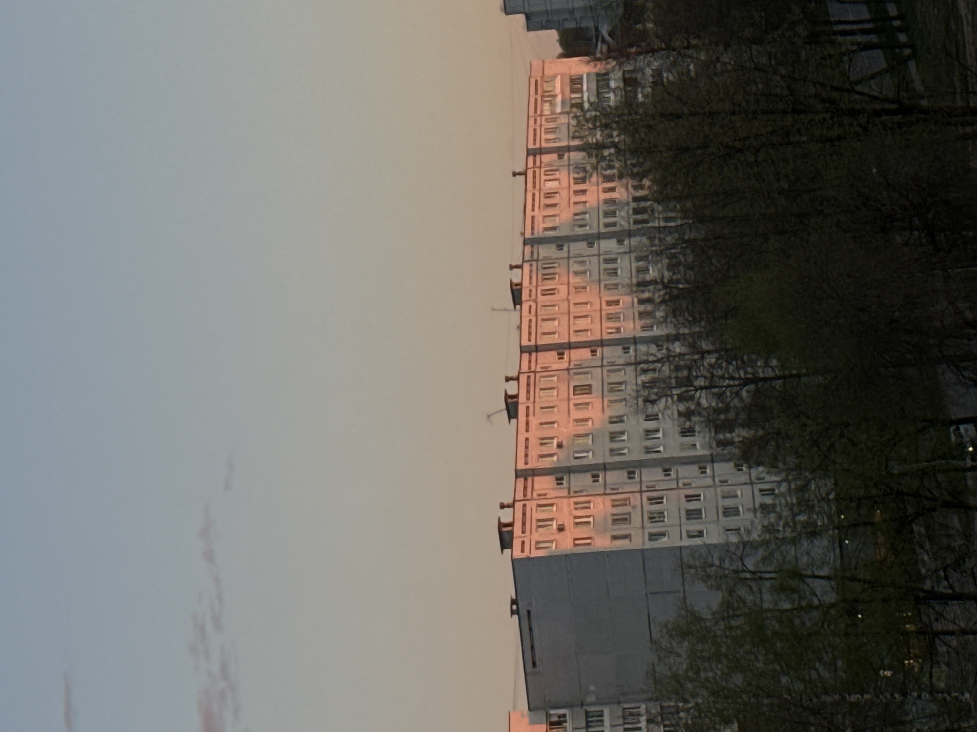 The image captures a high-rise apartment building in the city of Chaikovsky, Russia. The building is characterized by its repetitive grid of windows. The structure appears to be constructed from pre-fabricated concrete panels, a common feature of Soviet-era architecture. The building is situated in an urban environment, indicated by the presence of deciduous trees and possibly other buildings in the background. The presence of trees suggests a park or residential area. The sky is a gradient of light blue to a warm peach color, implying that the photo was taken either around sunrise or sunset. The lighting casts a soft, diffuse glow on the building's facade.