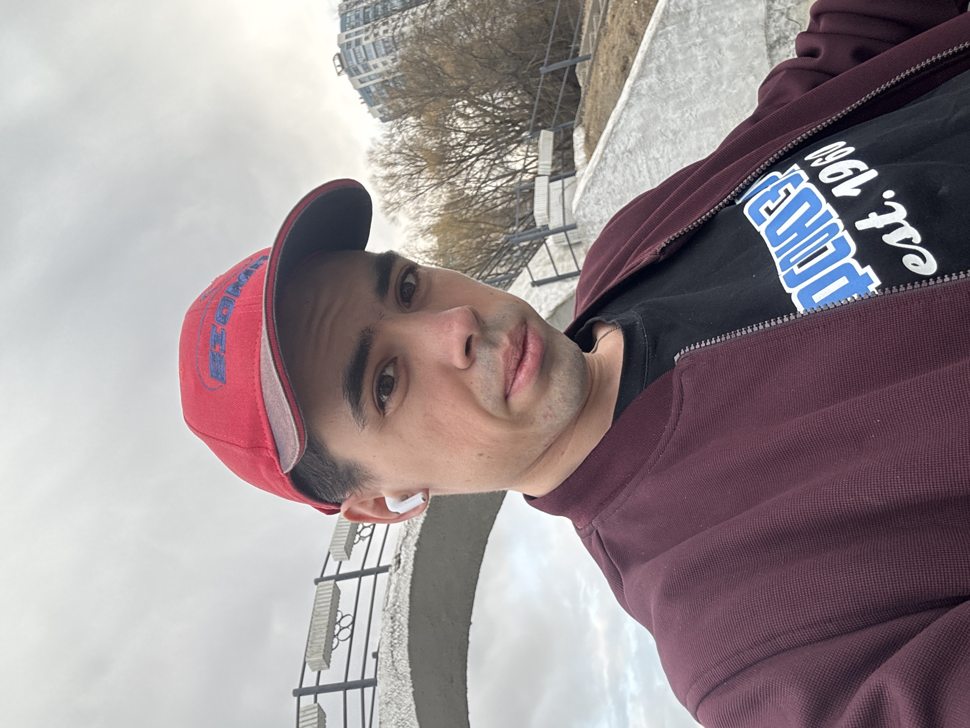 A man stands outdoors in Moscow, Russia on an overcast day, positioned near a concrete arched structure with metal railings. He wears a red baseball cap, a maroon zip-up jacket over a black t-shirt with "TECHNO est. 1960" printed on it, and an AirPod in his ear. The background includes a building with multiple floors and barren trees.