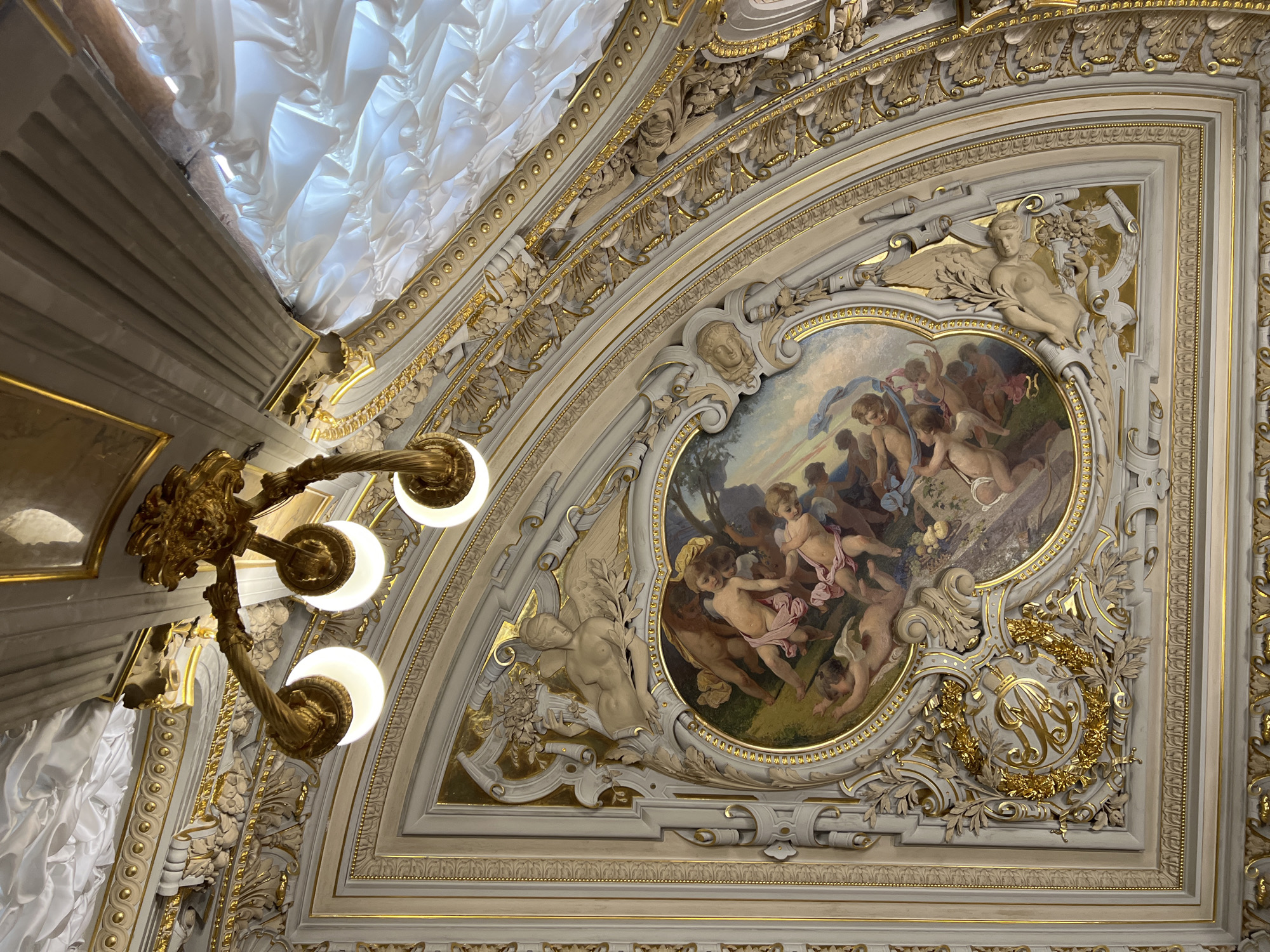 The image shows a close-up, low-angle view of an ornate ceiling in Saint Petersburg, Russia. The ceiling features a central oval painting depicting a group of cherubic figures in a pastoral landscape, possibly a mythological scene. The painting is framed by elaborate stucco decorations in gold and cream, including sculpted figures of winged beings, floral motifs, and a monogram within a golden wreath.
To the left, a gilded chandelier with three illuminated bulbs is visible, casting a warm glow on the decorative elements. The chandelier appears to be made of intricate metalwork, possibly brass or gold-plated.
The upper portion of the image reveals a section of the ceiling adorned with draped white fabric, creating a textured, voluminous effect that contrasts with the rigid architectural elements. This fabric could be part of the decorative scheme or a structural element.
The overall impression is one of opulence and grandeur, characteristic of historical palaces or theaters. The lighting suggests it is either daytime with artificial illumination or nighttime. The detailed artistry of the painting and stucco work, along with the gilded chandelier, indicates a high-end interior setting. No visible text is present in the image.