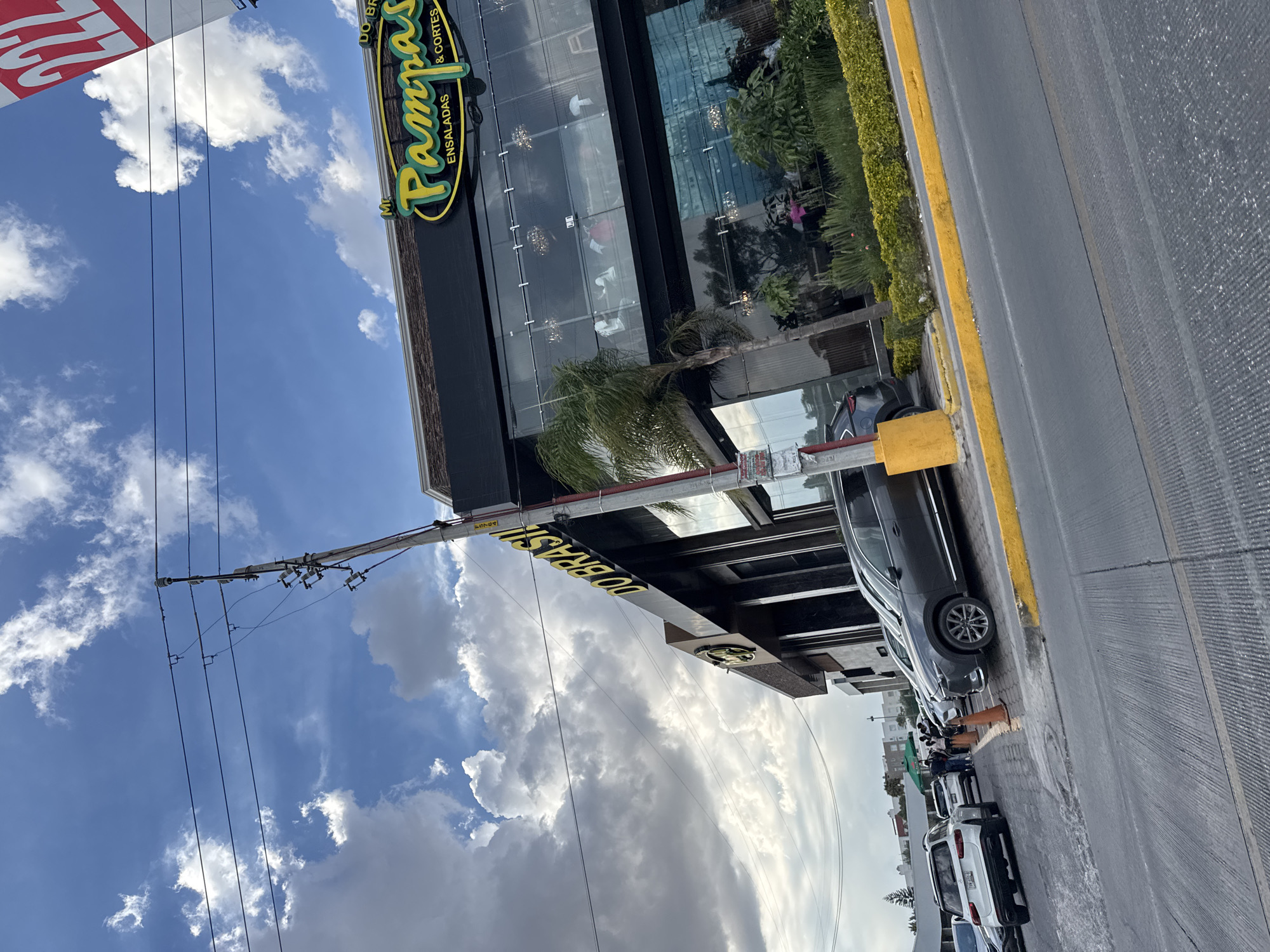 The image captures a street scene in San Bernardino Tlaxcalancingo, Mexico. Prominently featured is the building of the "Mr. Pampas" restaurant, identifiable by its yellow and green logo. The building has a modern glass facade. Cars are parked along the street adjacent to the building, with a yellow painted curb line marking the edge of the road. Overhead, power lines traverse the sky, which is filled with clouds. A secondary sign “DO BRASIL” is also visible. The scene appears to be during daylight hours.