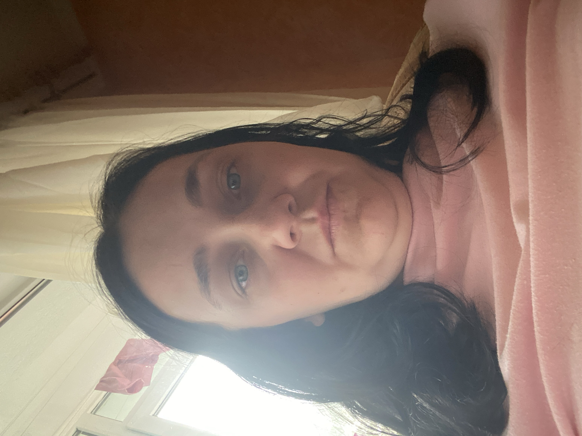 A medium shot shows a woman from the chest up, facing the camera. She has dark hair, blue eyes, and is wearing a pink top. The background consists of a window with sheer curtains on the left and a brown wall on the right. A pink piece of fabric is hanging from the window frame. The lighting suggests an indoor setting, possibly a home. The woman's expression is neutral. The scene is static and does not show any particular action or interaction. The location is unspecified.