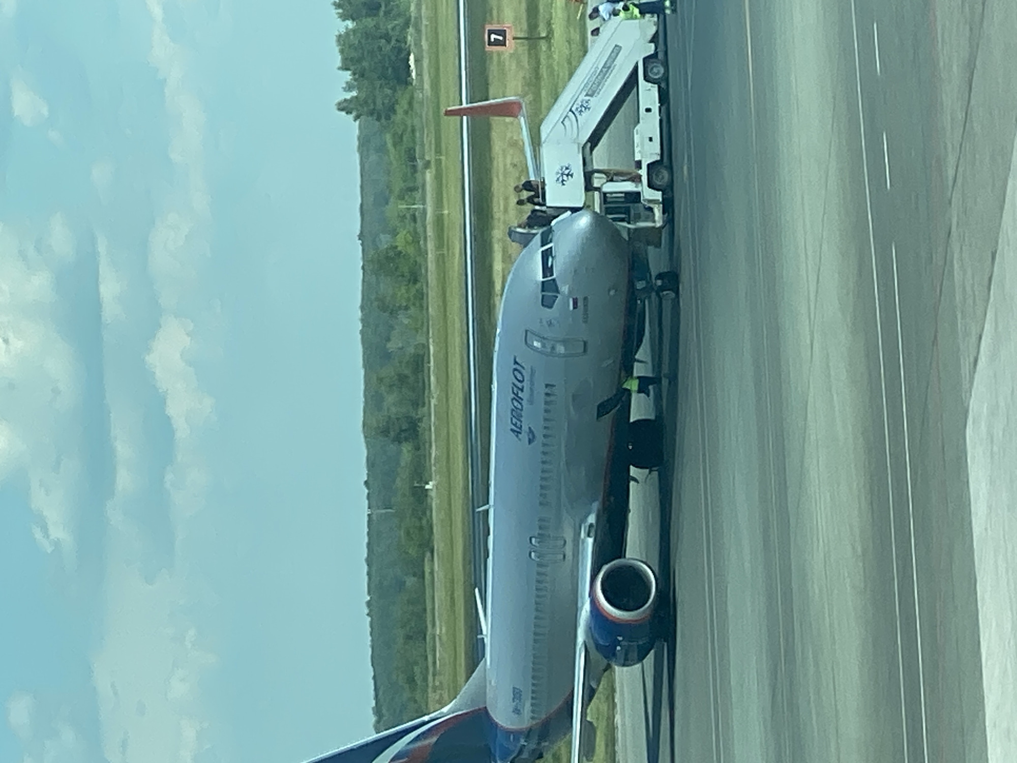 The image shows an Aeroflot airplane parked on the tarmac at Tolmachevo Airport (OVB) in Ob, Russia. The aircraft, a Boeing 737, is positioned at Gate 7, indicated by a sign on a pole to the right of the plane. A mobile staircase is connected to the front left door of the aircraft, and several people, likely passengers and ground crew, are visible near the entrance. The sky is partly cloudy, and a line of trees and foliage forms the background. The scene suggests routine airport operations, with the plane prepared for boarding or disembarking.