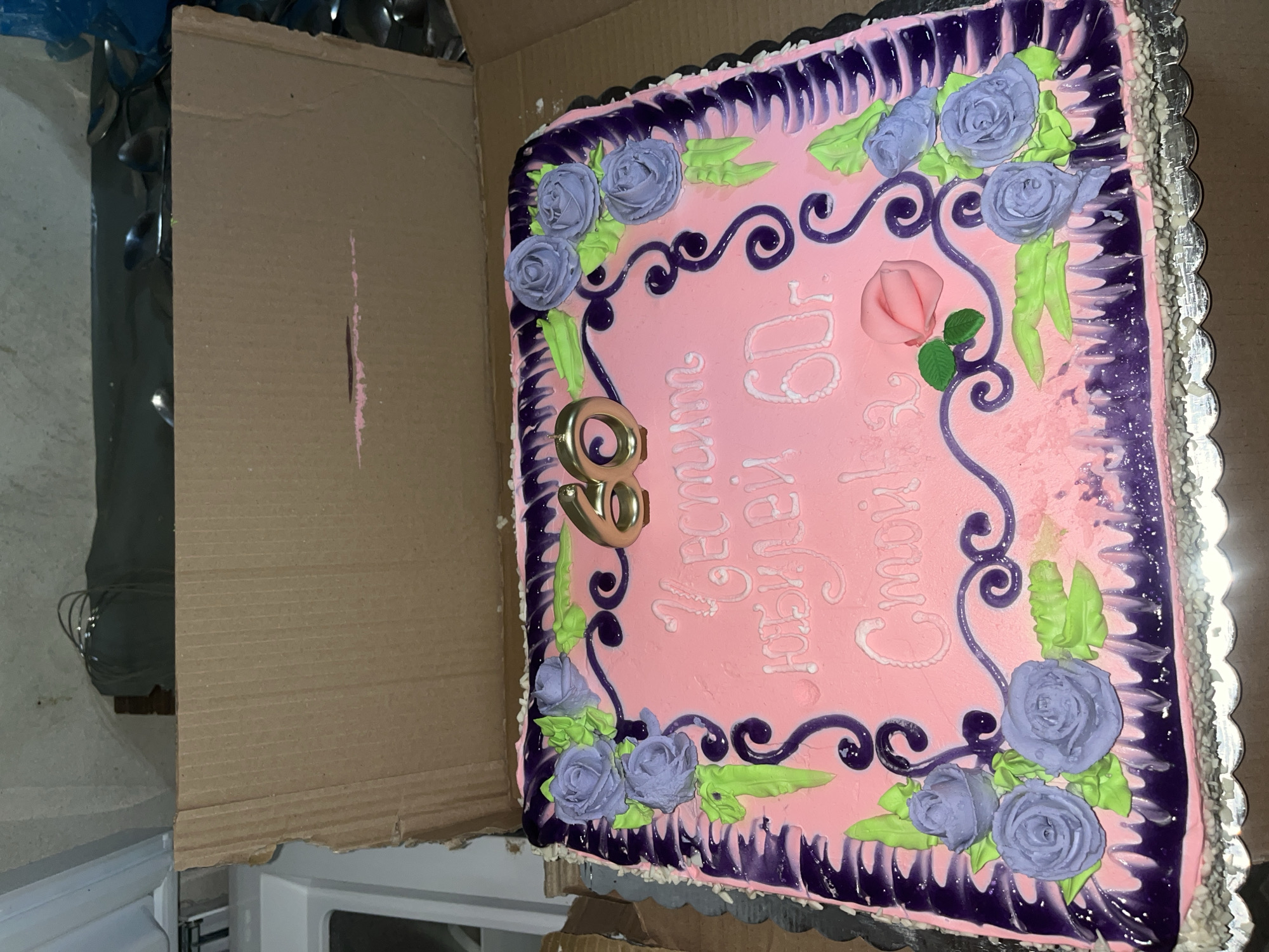 The image shows a decorated square cake inside a cardboard box. The cake has pink frosting, with violet roses, green leaves, and purple swirls. White lettering spells out a message in what appears to be Bulgarian, accompanied by a rose and the golden number "60". A silver scalloped edge surrounds the cake. The scene is likely indoors, but the specific location in Kosten, Bulgaria cannot be determined from the image.